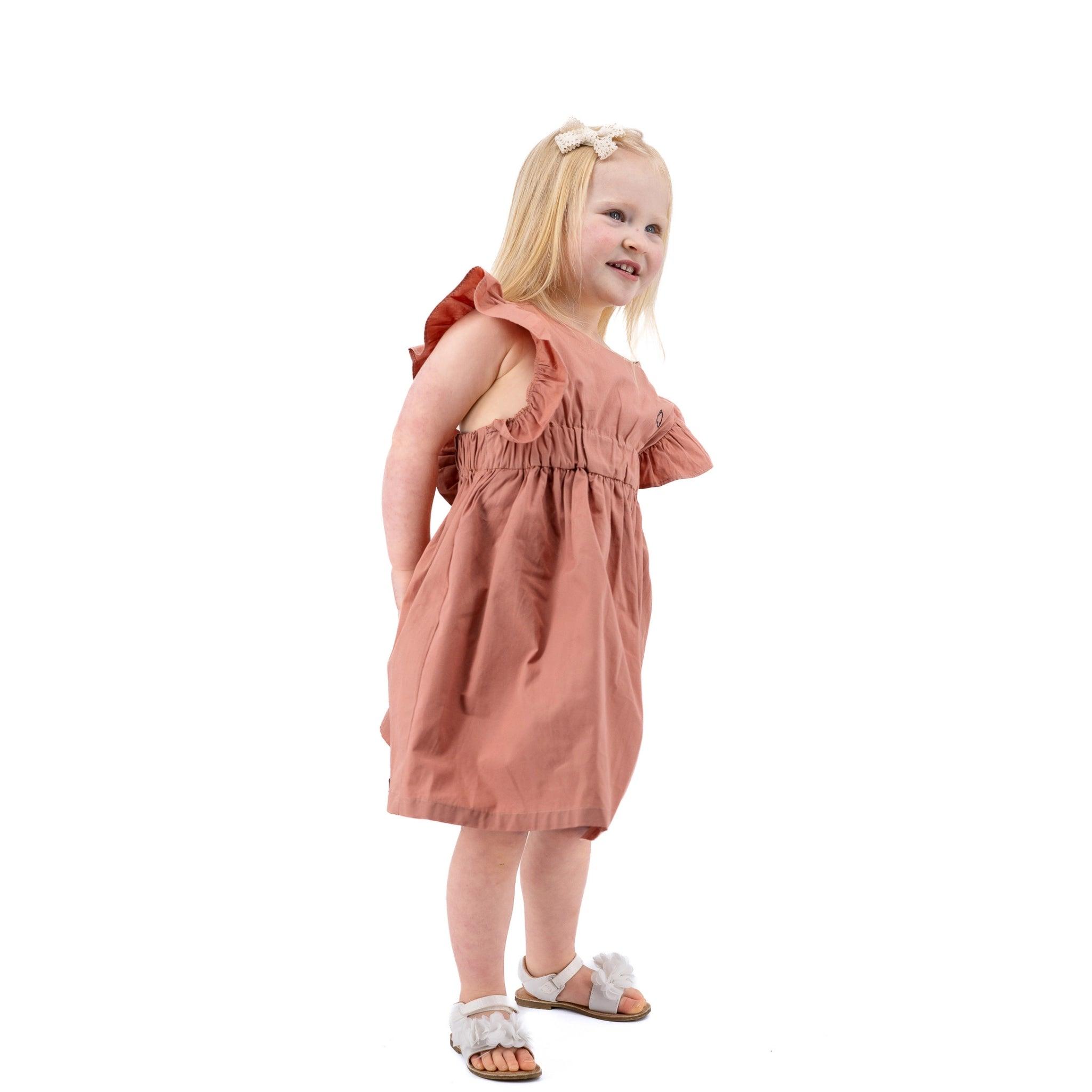 A young girl with blonde hair, wearing a ruffled Karee brick dust cotton floral dress and white sandals, smiling and looking over her shoulder against a white background.