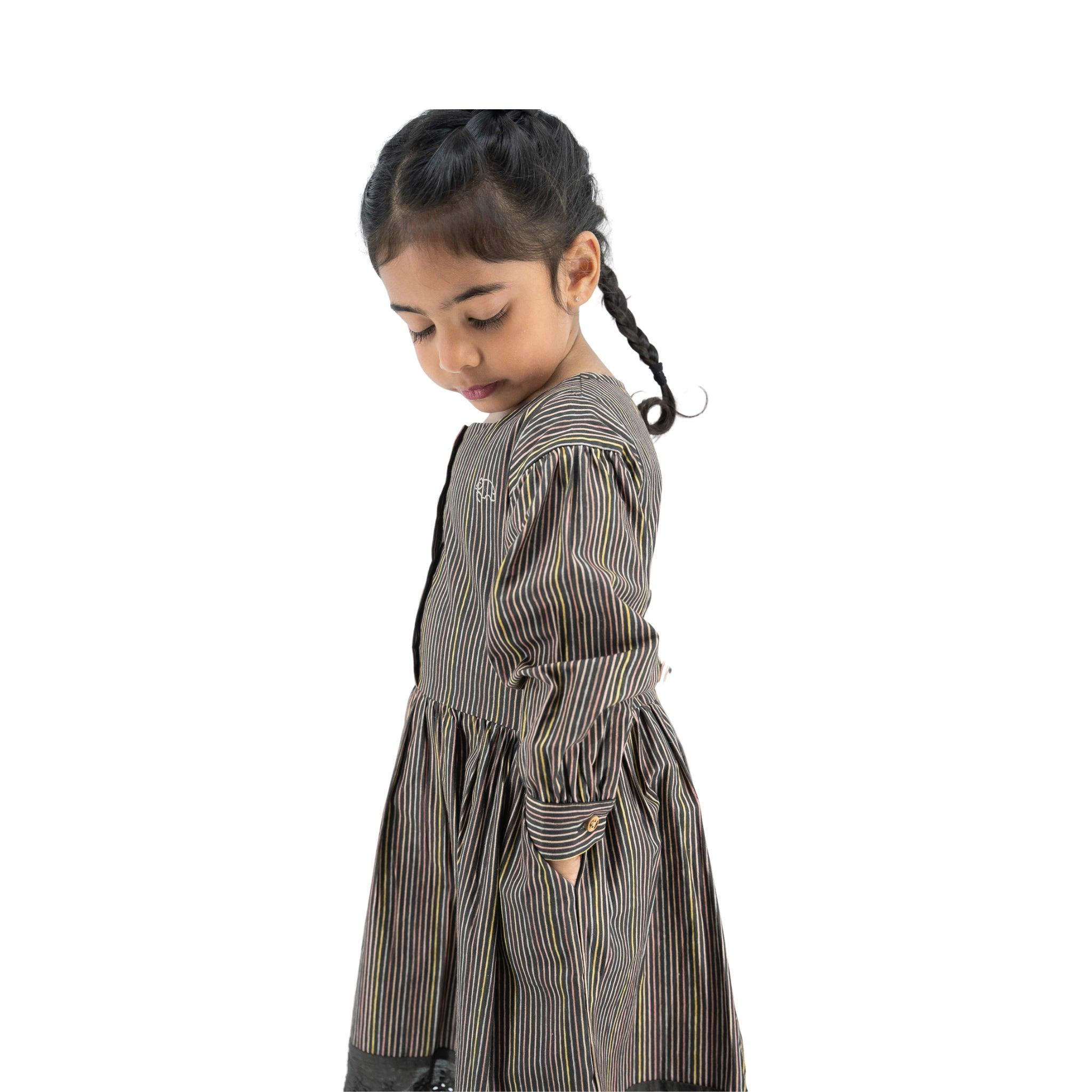 Young girl with braided hair, wearing a Karee black striped full sleeve cotton dress, standing sideways and looking down, isolated on a white background.