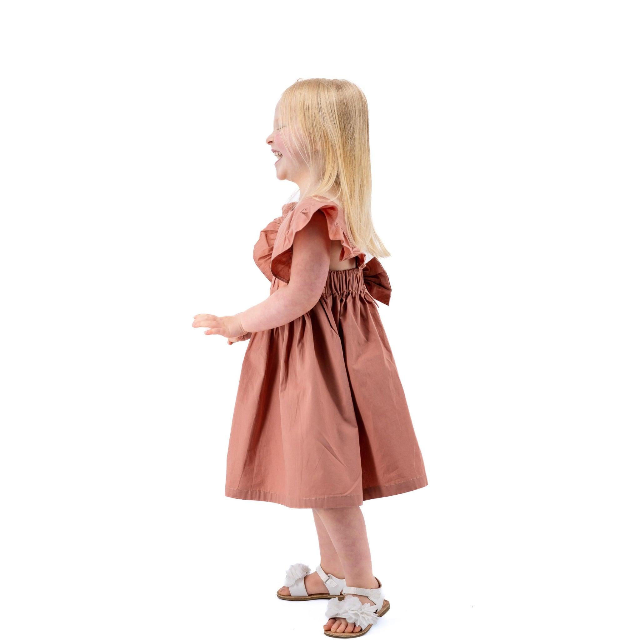 Young girl in a Karee Brick Dust Cotton Floral Dress for Girls and white sandals smiling and walking to her right on a white background.