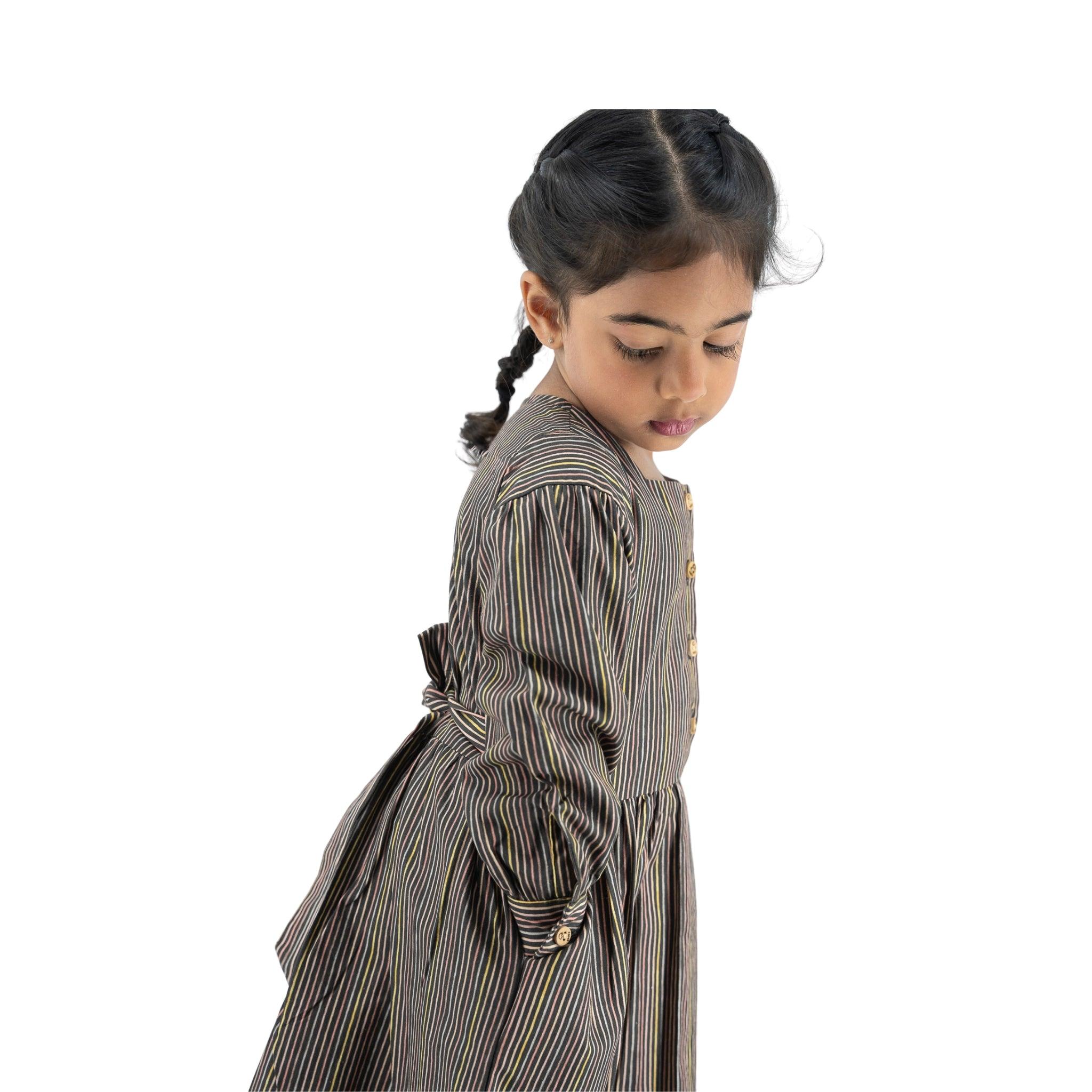 Young girl with braided hair, wearing a Karee Black Striped Full sleeve Cotton Dress, looking downward to her left side, isolated on white background.