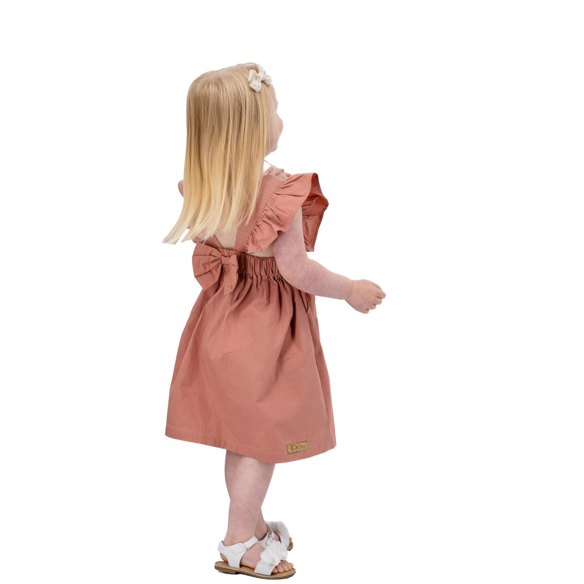 Young girl with long blonde hair wearing a Karee Brick Dust Cotton Floral Dress for Girls and white sandals, standing and looking to the side, isolated on a white background.