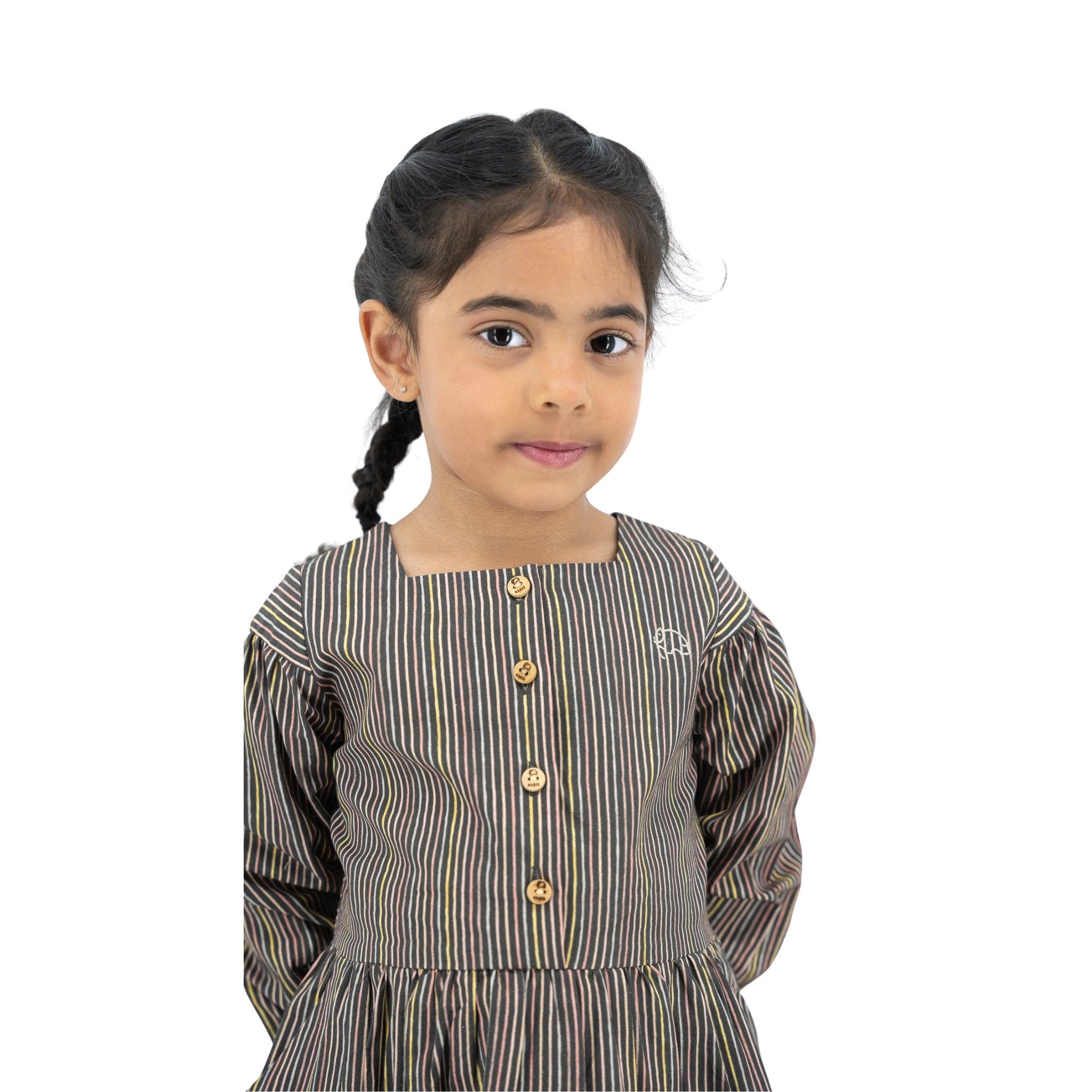 Young girl with dark hair in braids wearing a Karee black striped full sleeve cotton dress, standing against a white background.