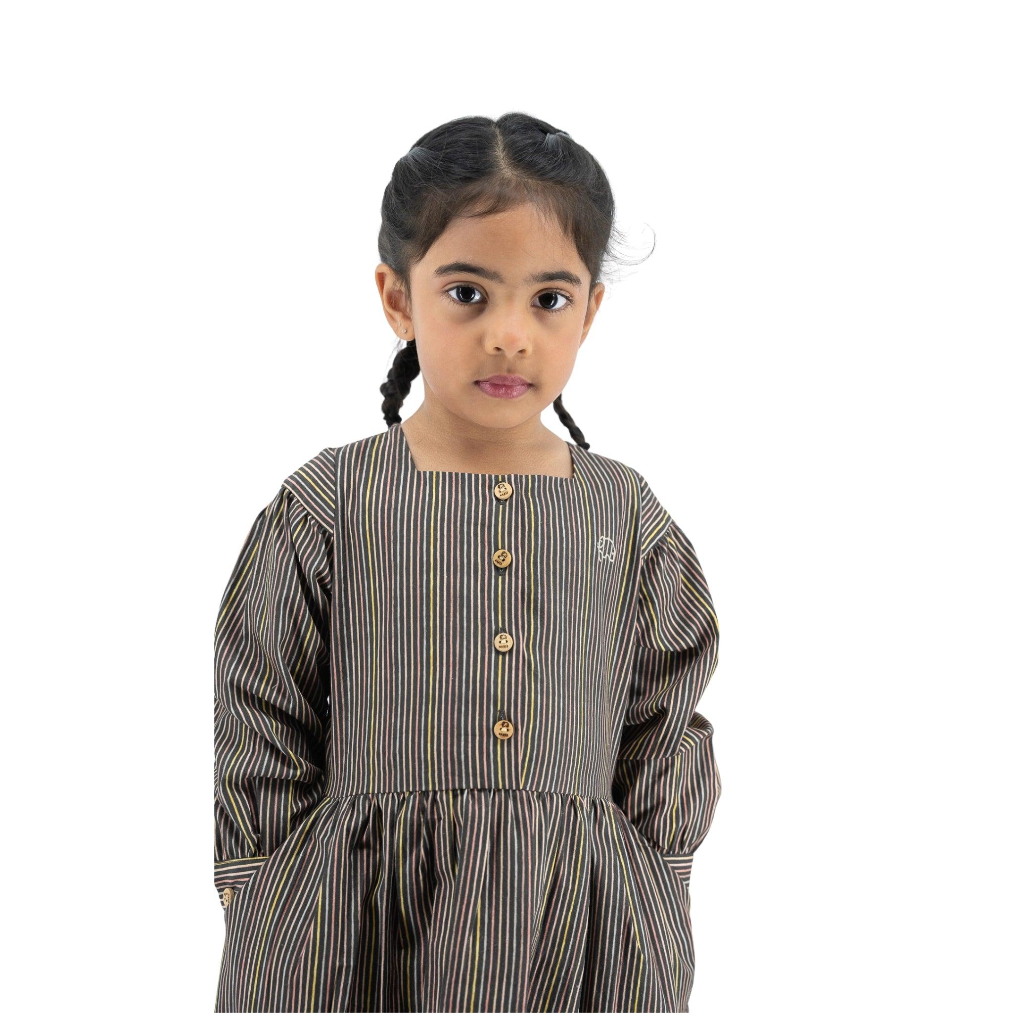 Young girl with braided hair wearing a Karee Black Striped Full sleeve Cotton Dress, standing against a white background, looking slightly to the side.