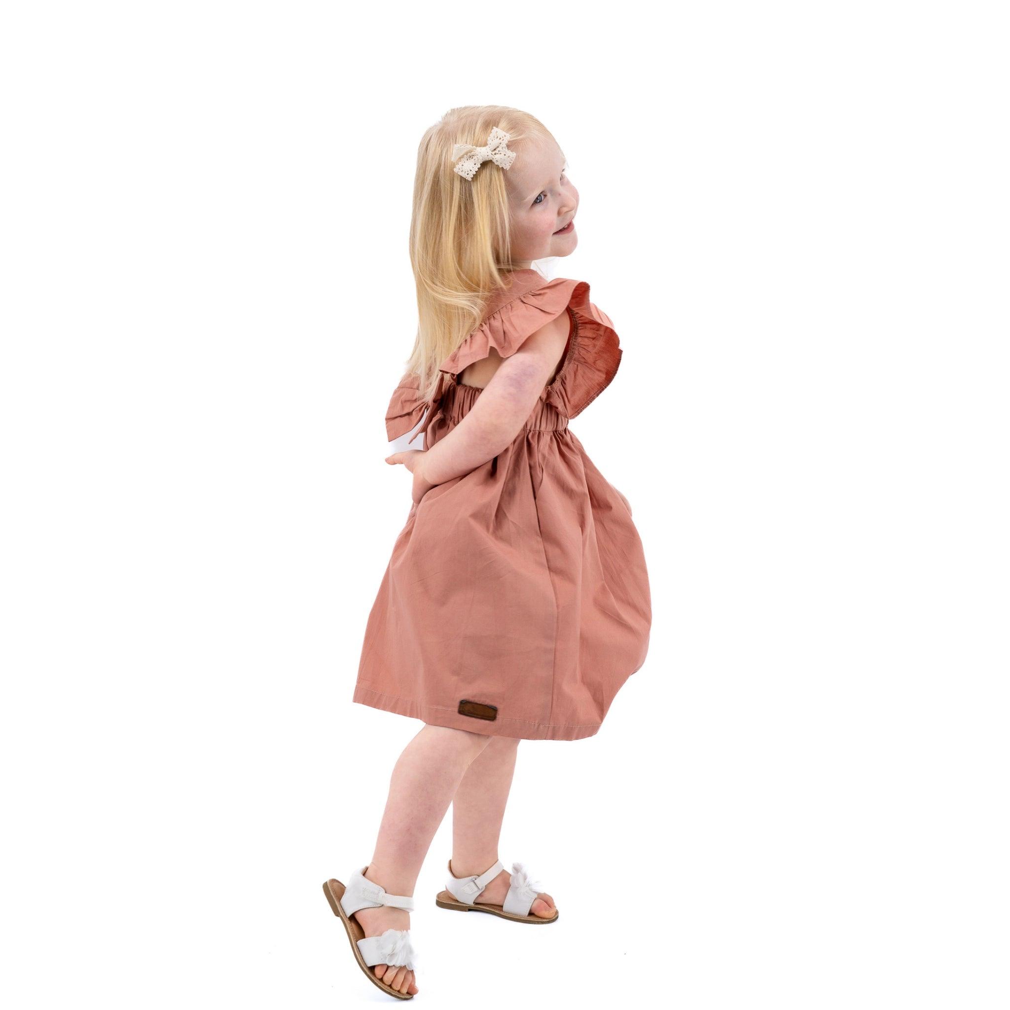 A young girl with blonde hair, wearing a Karee Brick Dust Cotton Floral Dress for Girls and white sandals, looks over her shoulder with a smile, standing against a white background.