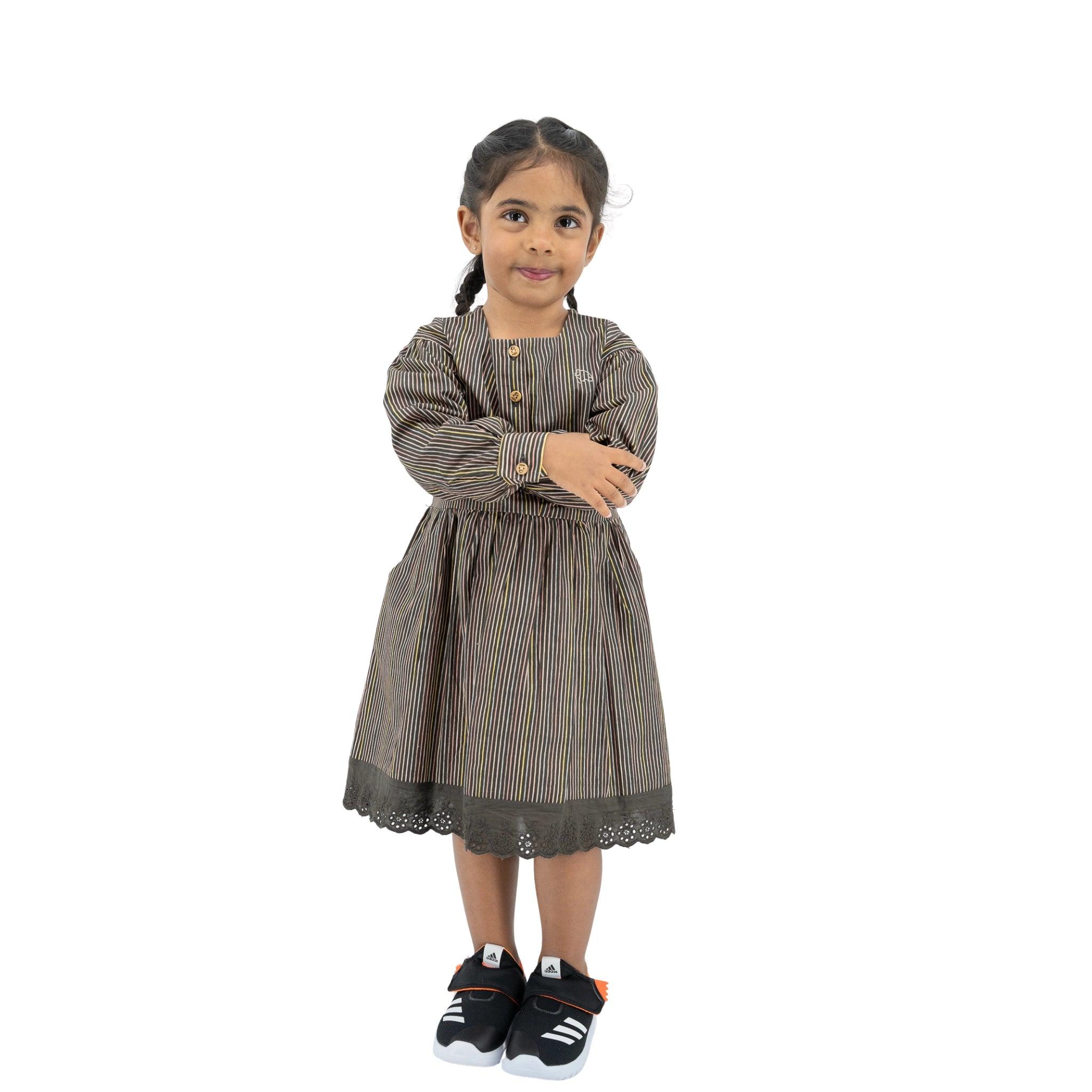 Young girl standing with arms crossed, wearing a Karee black striped full sleeve cotton dress and sneakers, smiling at the camera on a white background.