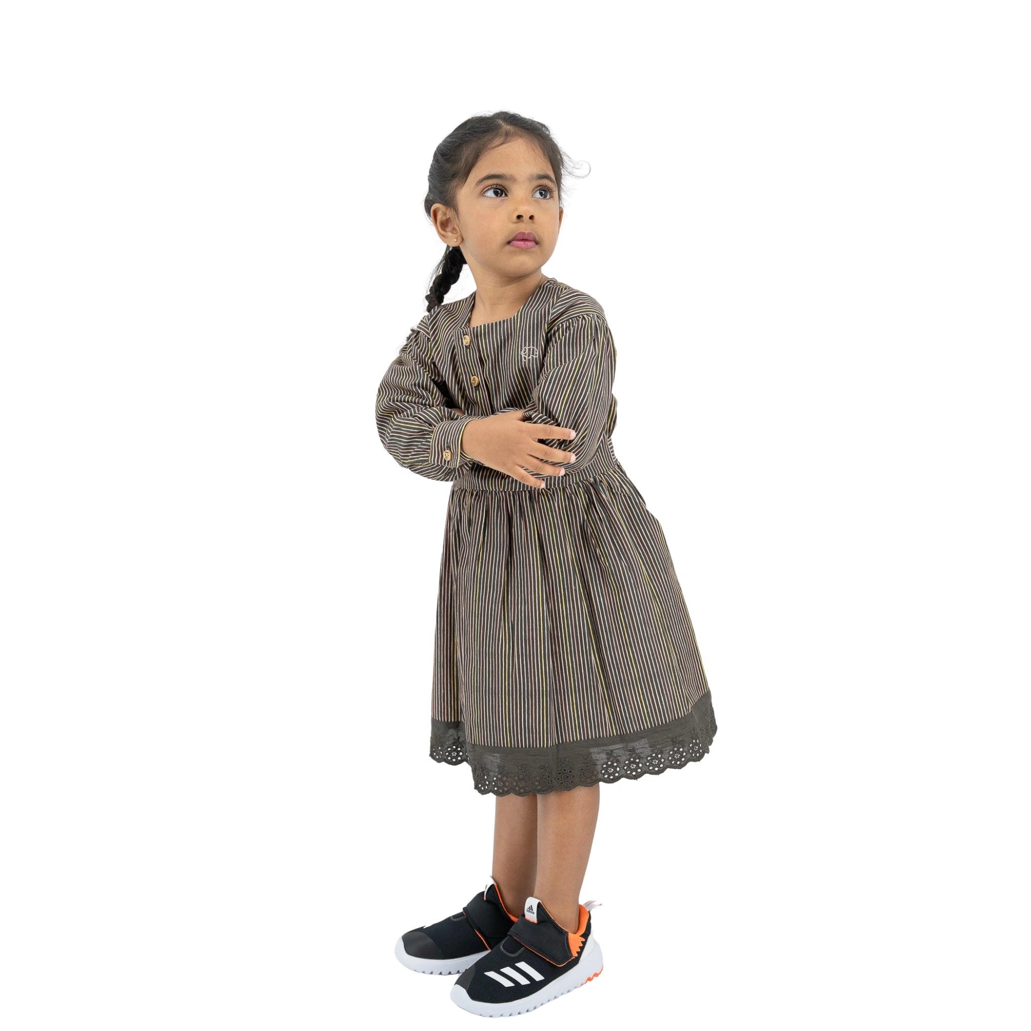 Young girl standing, wearing a Karee Black Striped Full sleeve Cotton Dress and sneakers, looking upwards to the left, isolated on a white background.