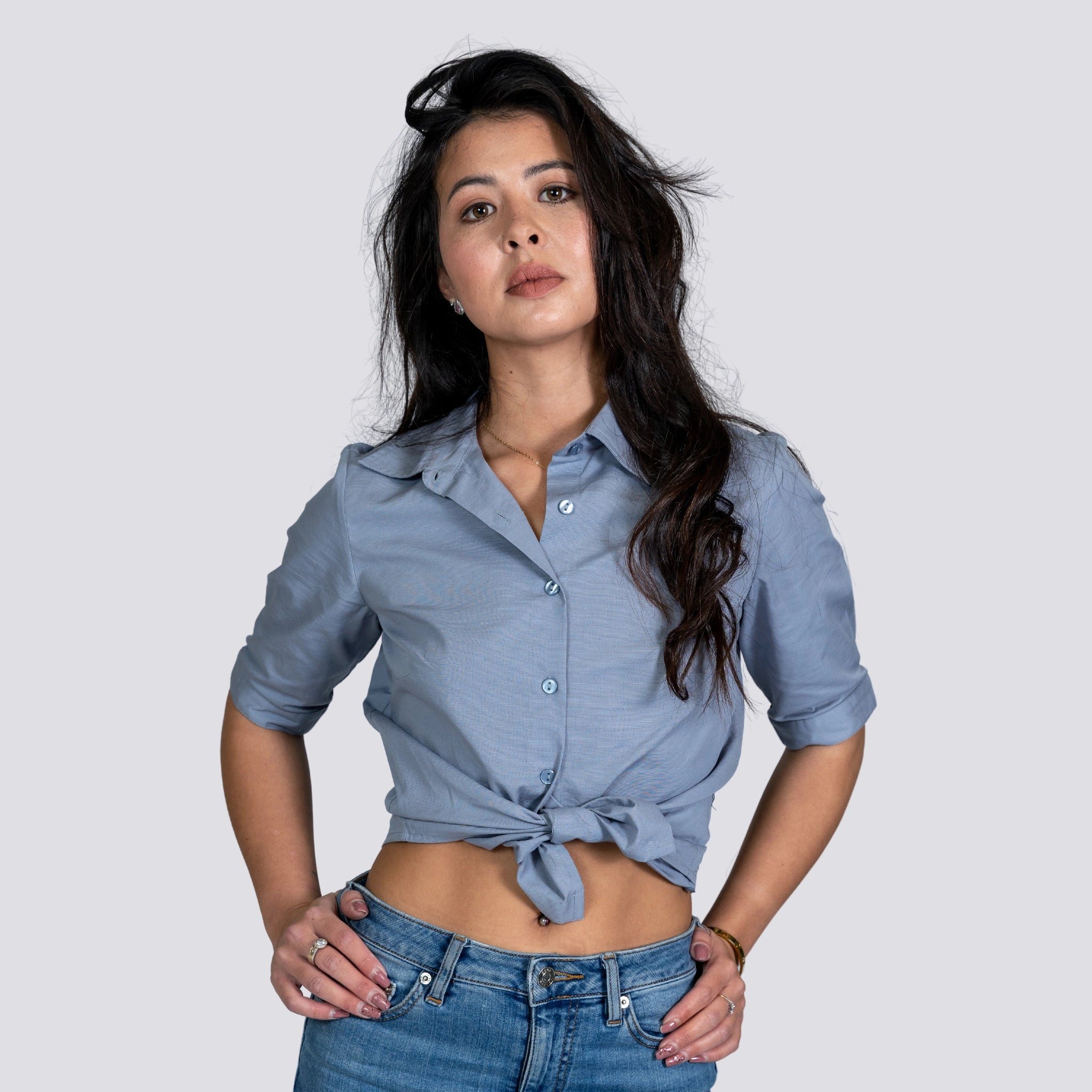 A woman with long dark hair wearing a Karee Grey Mist Linen Shirt For Women and jeans, standing confidently against a grey background.
