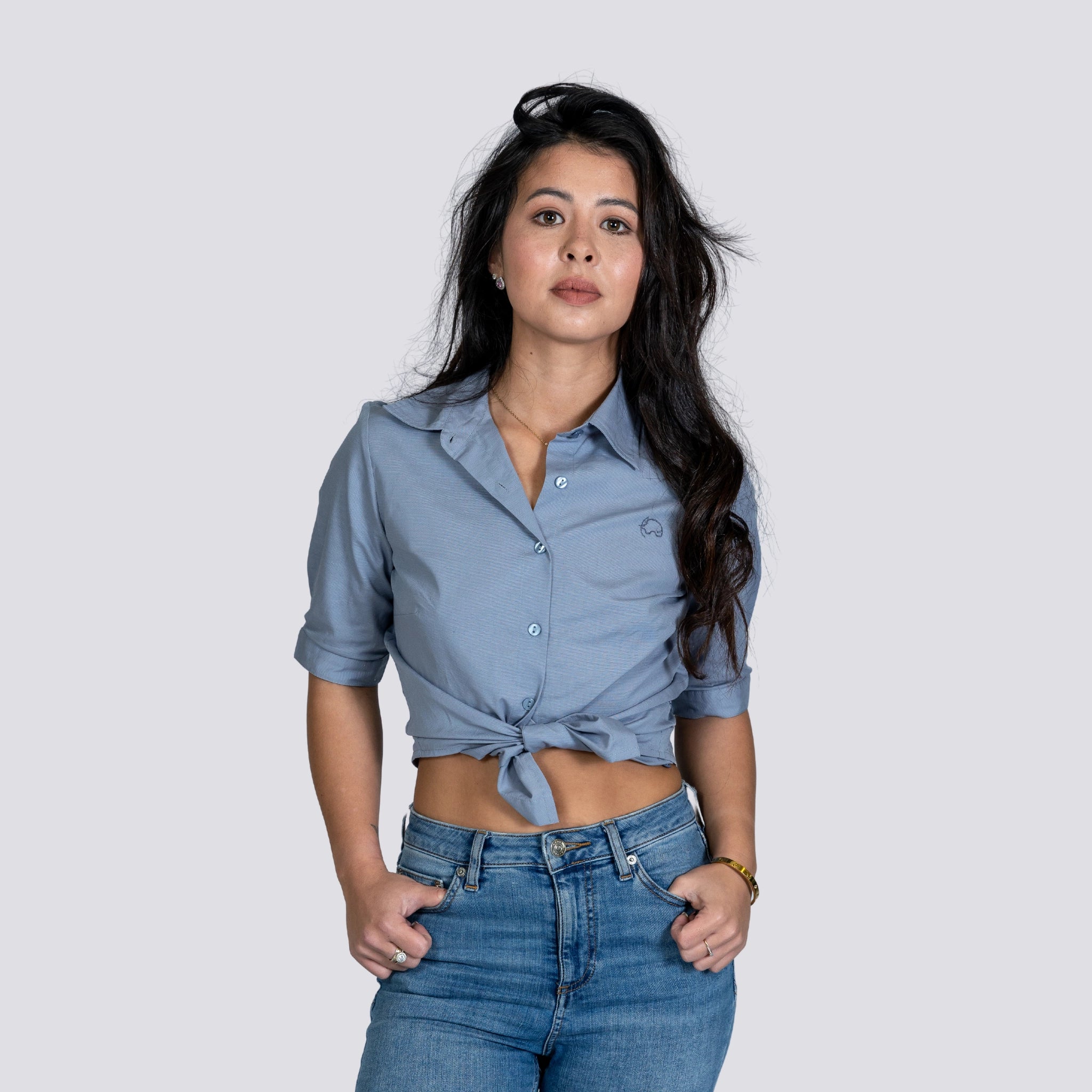 A young woman with long black hair wearing a versatile essential, Grey Mist Linen Shirt For Women by Karee, and jeans stands against a gray background.