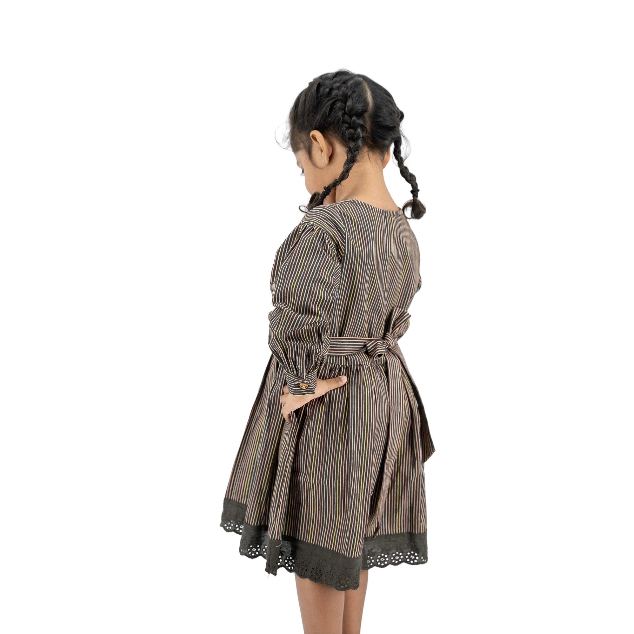 A young girl in a Karee Black Striped Full sleeve Cotton Dress with a waist tie and lace trim, seen from the back, standing against a white background.