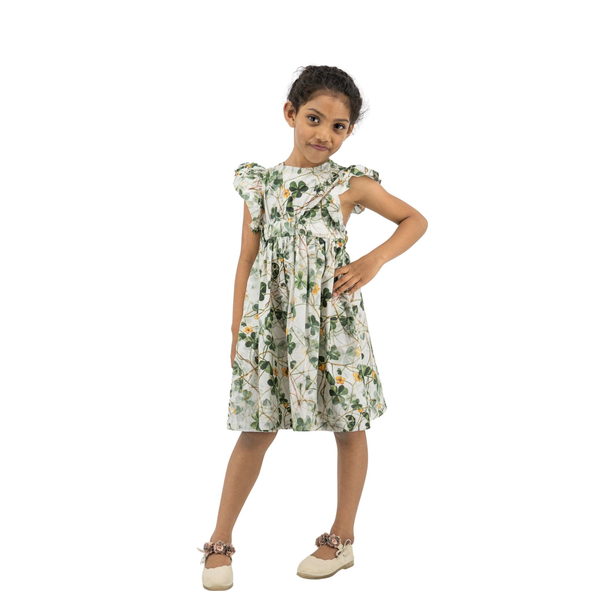 Young girl posing confidently, wearing a Karee green floral cotton dress and light brown shoes, standing against a white background.