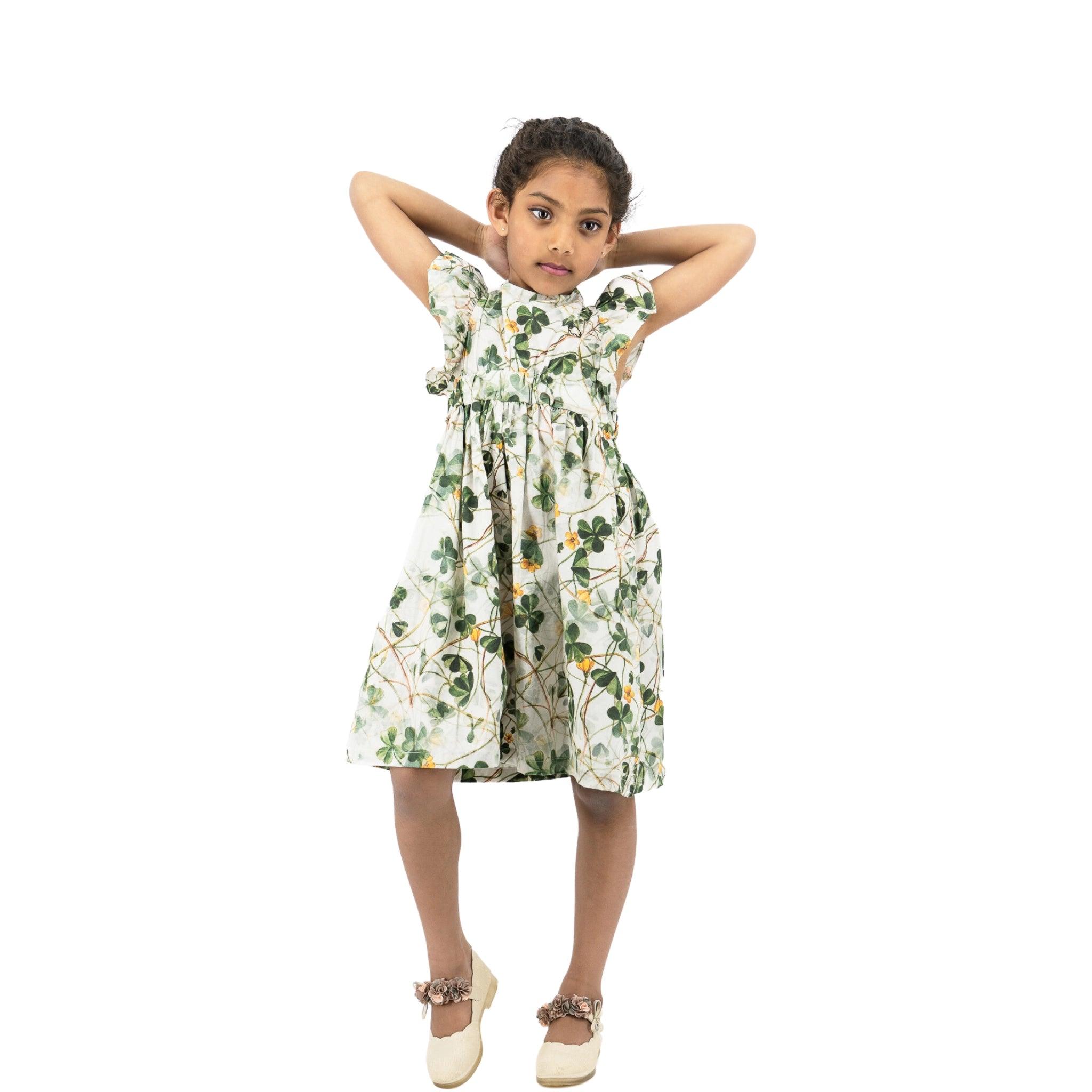 Young girl in a Karee green floral cotton dress standing with her hands behind her head, looking at the camera, against a white background.