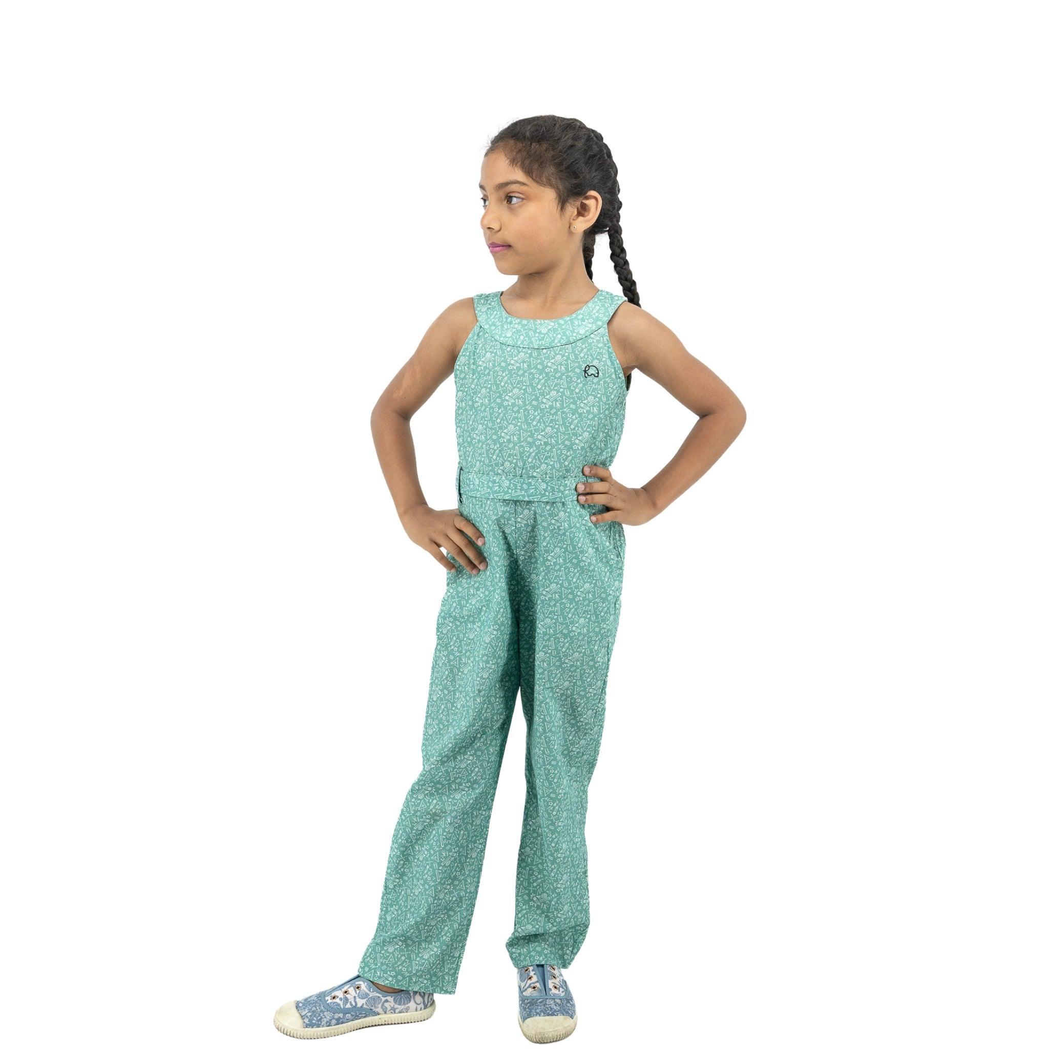 A young girl stands confidently, hands on hips, wearing a Karee Smoke Green Cotton Jumpsuit for Girls and patterned sneakers. She has long braided hair.