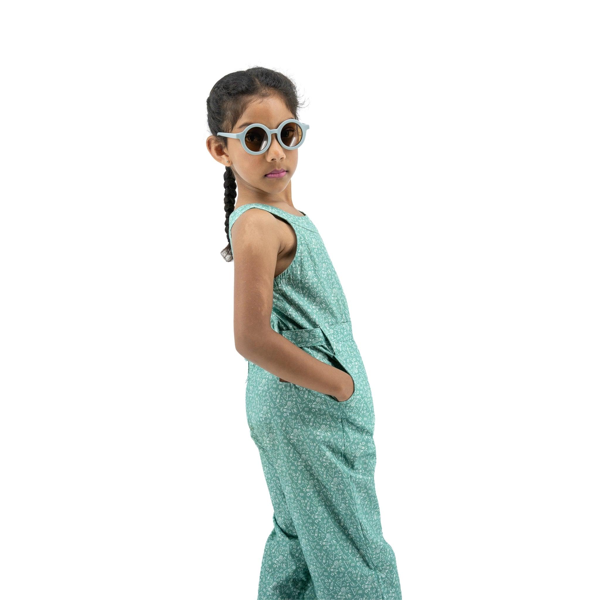 Young girl wearing eco-friendly sunglasses and a Karee Smoke Green Cotton Jumpsuit For Girls, standing sideways with hands on hips, isolated on a white background.