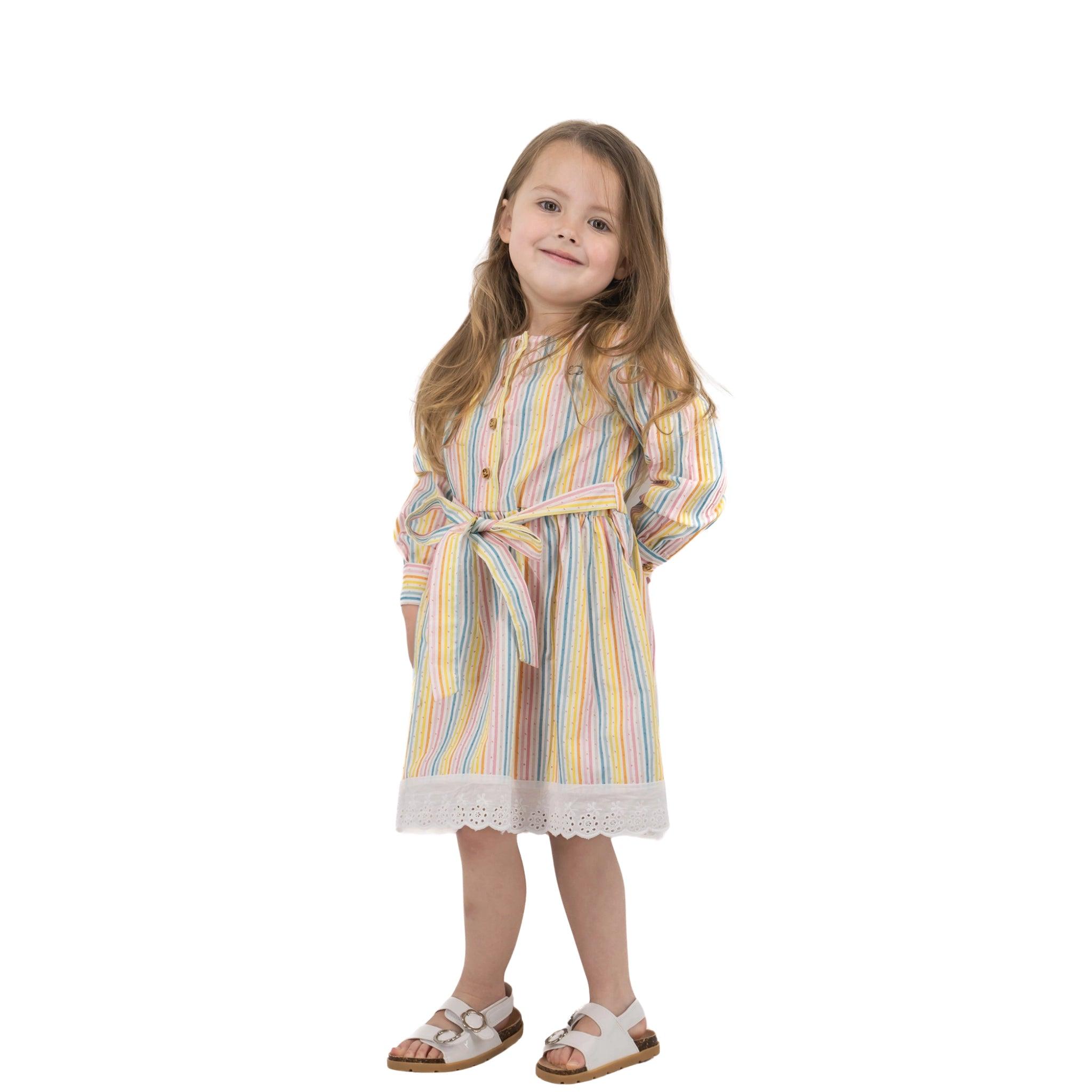 A young girl in a Karee striped pastel Long Puff Sleeve White Cotton Dress with Stripes and white sandals smiling and posing against a white background.