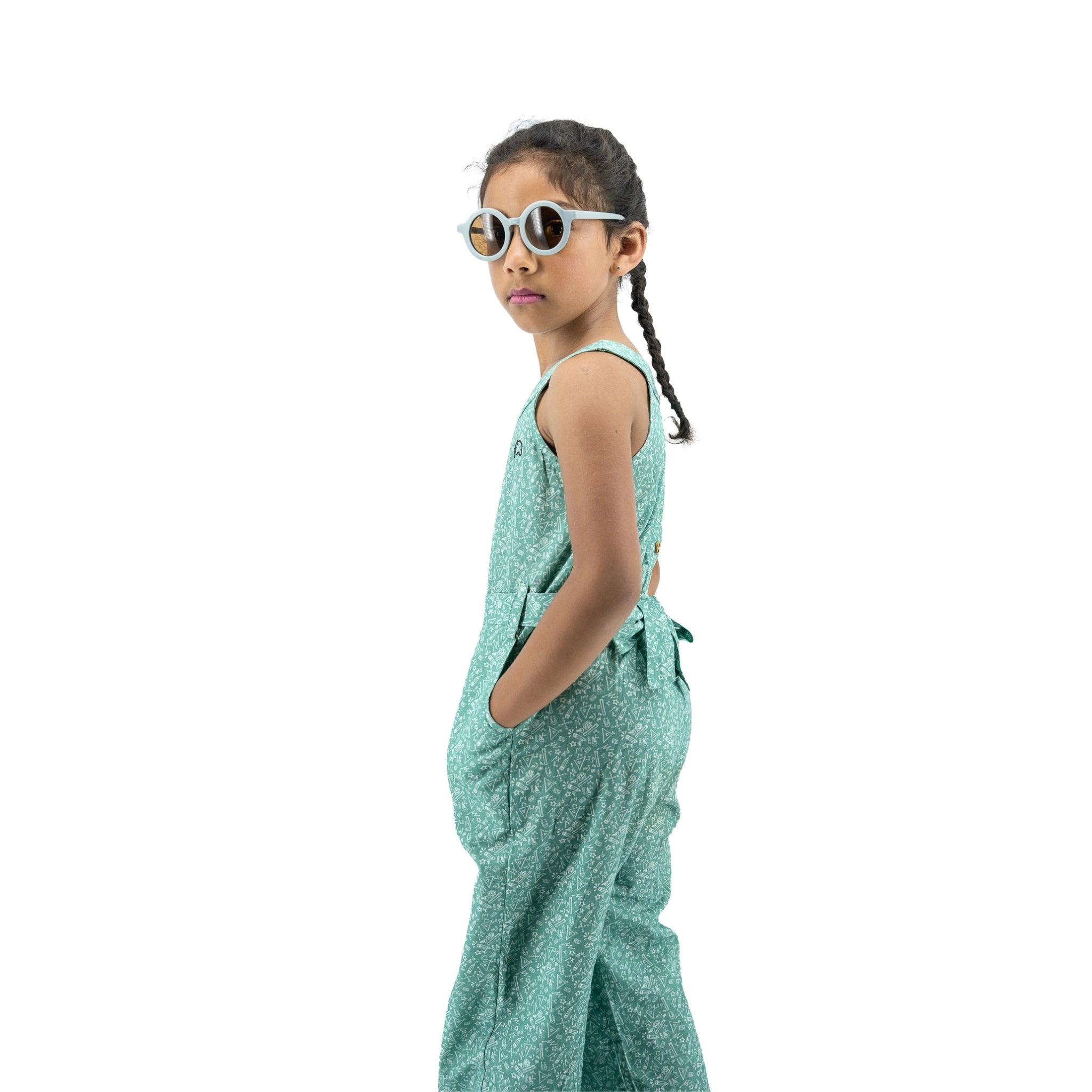A young girl with braided hair wearing sunglasses and a Smoke Green Cotton Jumpsuit For Girls by Karee stands in profile, looking over her shoulder against a white background.