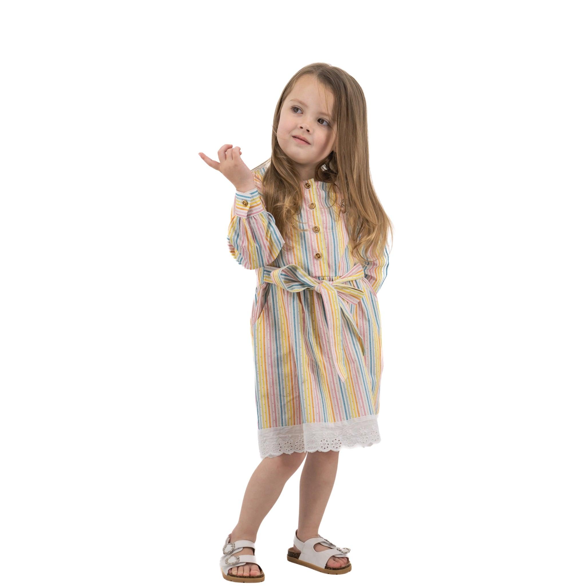 A young girl wearing a Karee Long Puff Sleeve White Cotton Dress with Stripes and sandals gestures with one hand, standing against a white background.