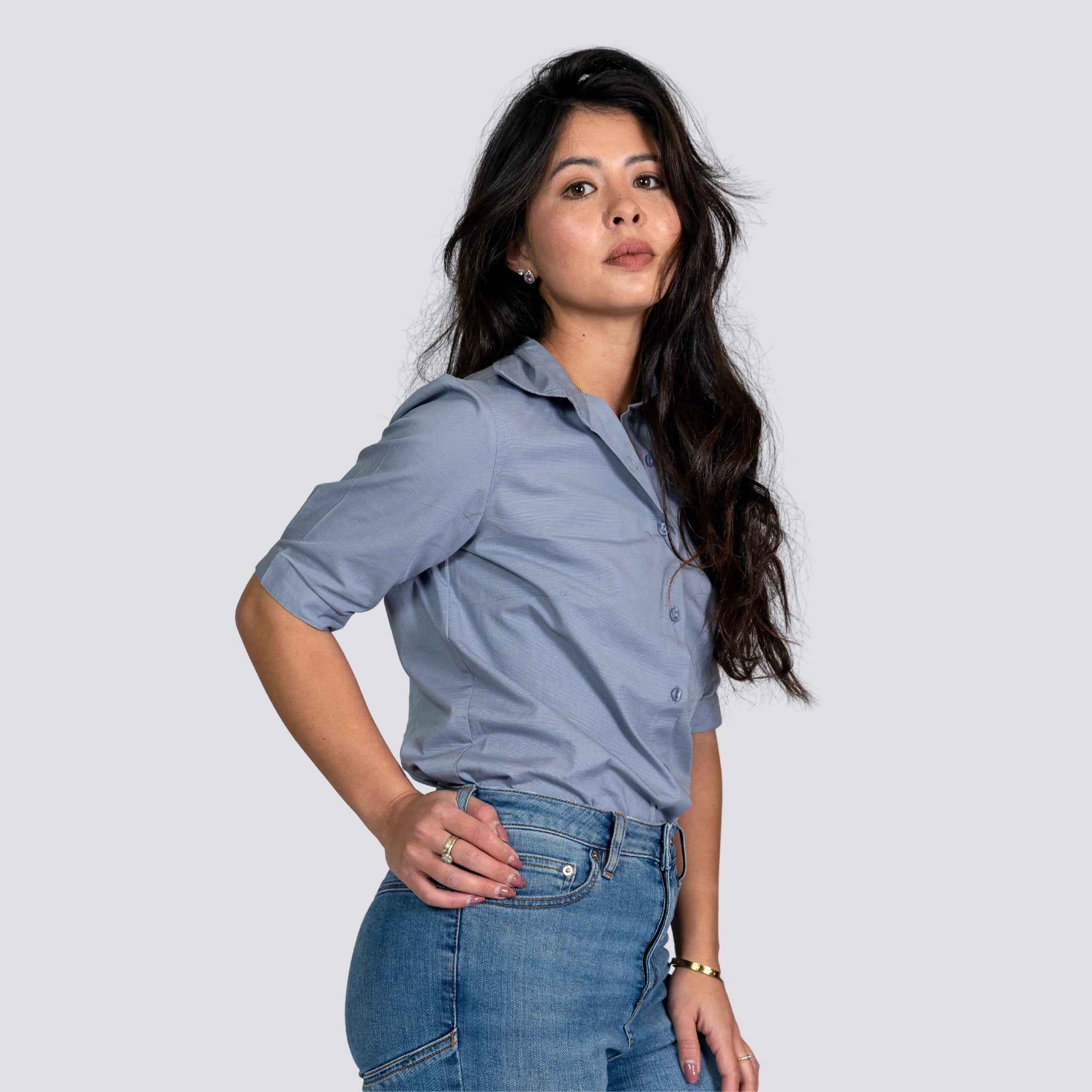 A woman with long dark hair wearing a Karee Grey Mist Linen Shirt For Women and blue jeans, posing with one hand on her hip against a light gray background.