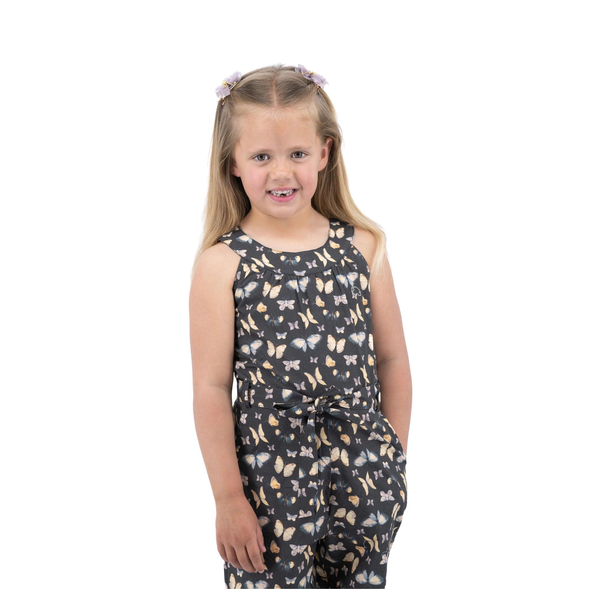 A young girl with floral hair accessories, smiling in a sleeveless butterfly print jumpsuit with a waist belt, standing against a white background. She is wearing the Karee Pirate Black Adventure-ready Cotton Play Suit.
