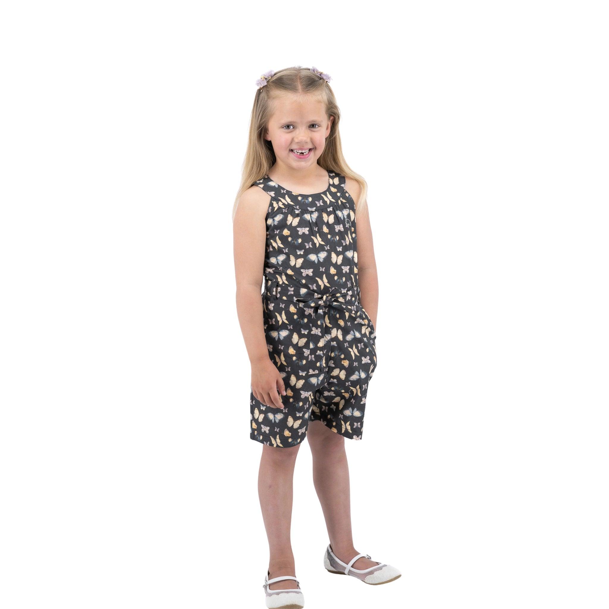 A young girl in a Karee Pirate Black Adventure-ready Cotton Play Suit with a waist belt and white shoes smiling and posing against a white background.