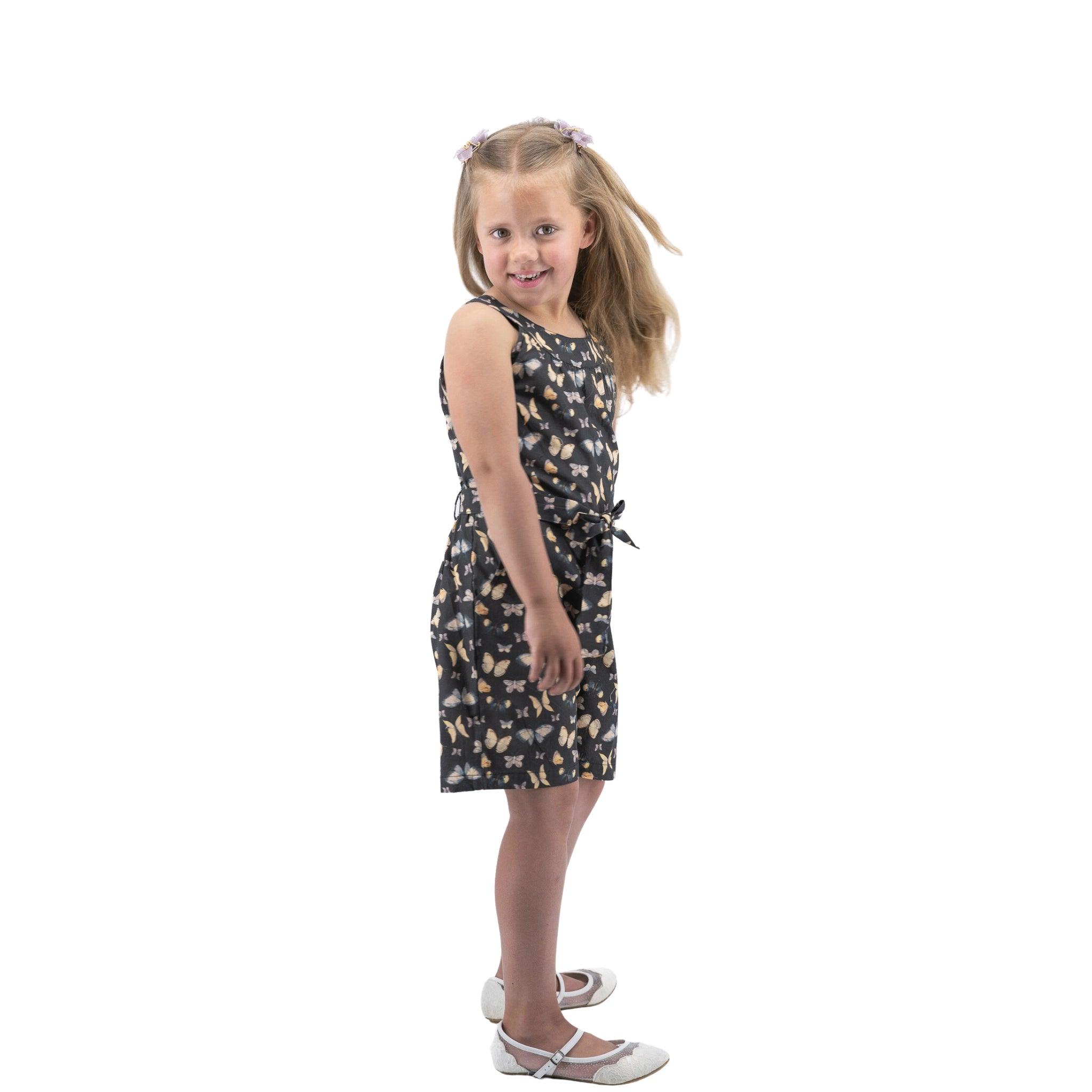Young girl in a sleeveless floral dress and white shoes smiling over her shoulder, standing against a white background wearing the Karee Pirate Black Adventure-ready Cotton Play Suit.