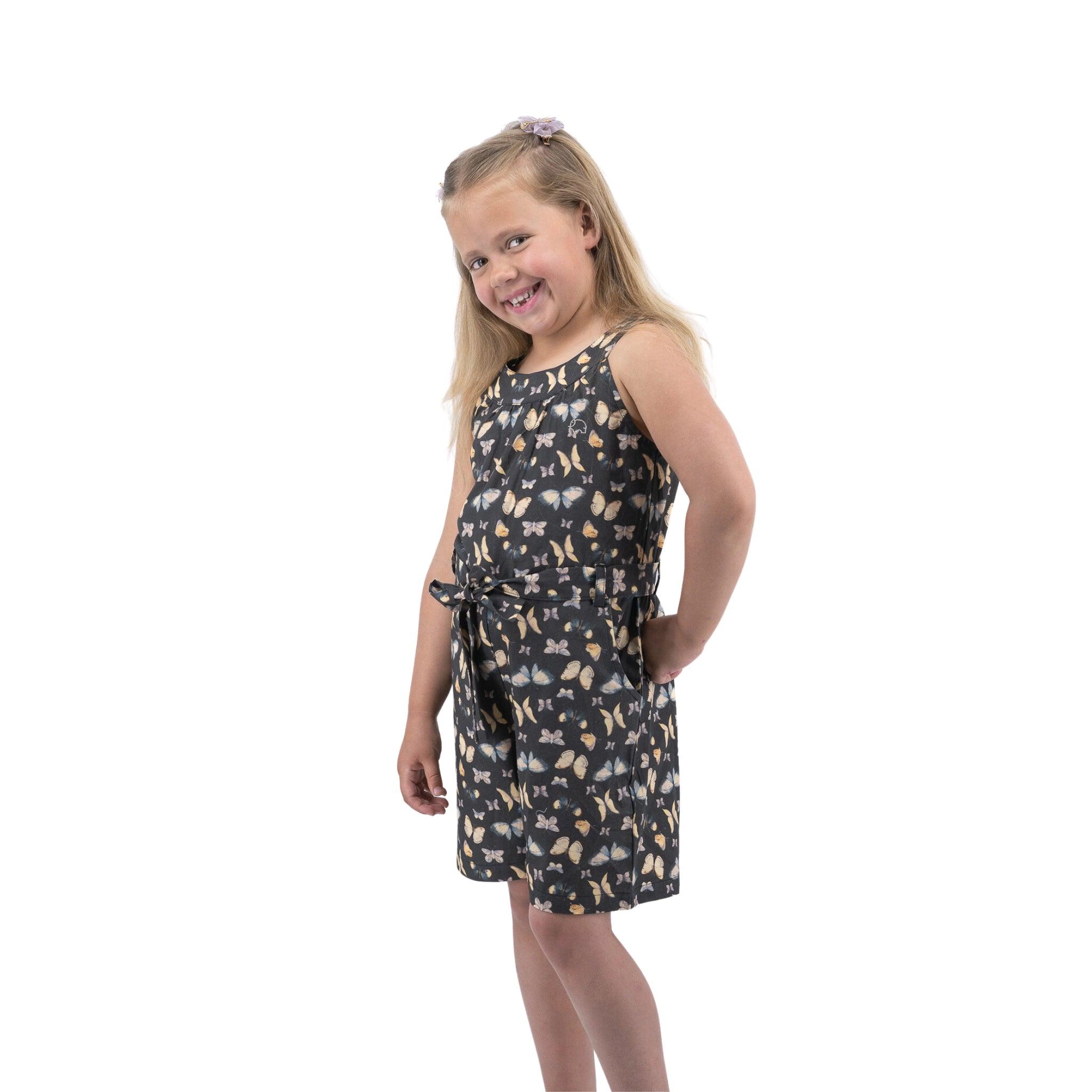 A young girl smiling at the camera, wearing a sleeveless floral dress, standing against a white background.