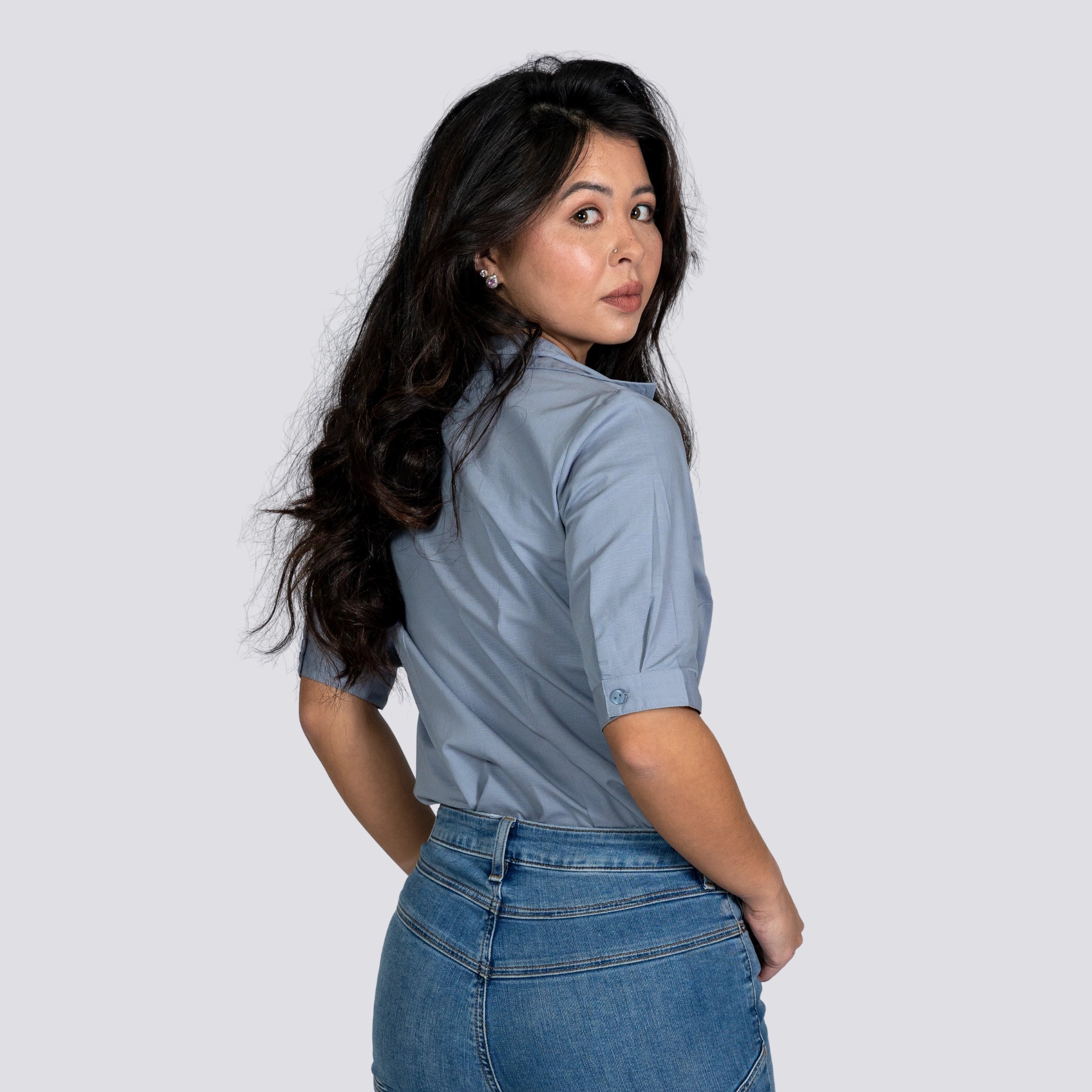 Asian woman in Karee Grey Mist Linen Shirt For Women and jeans, looking over her shoulder, standing against a gray background.