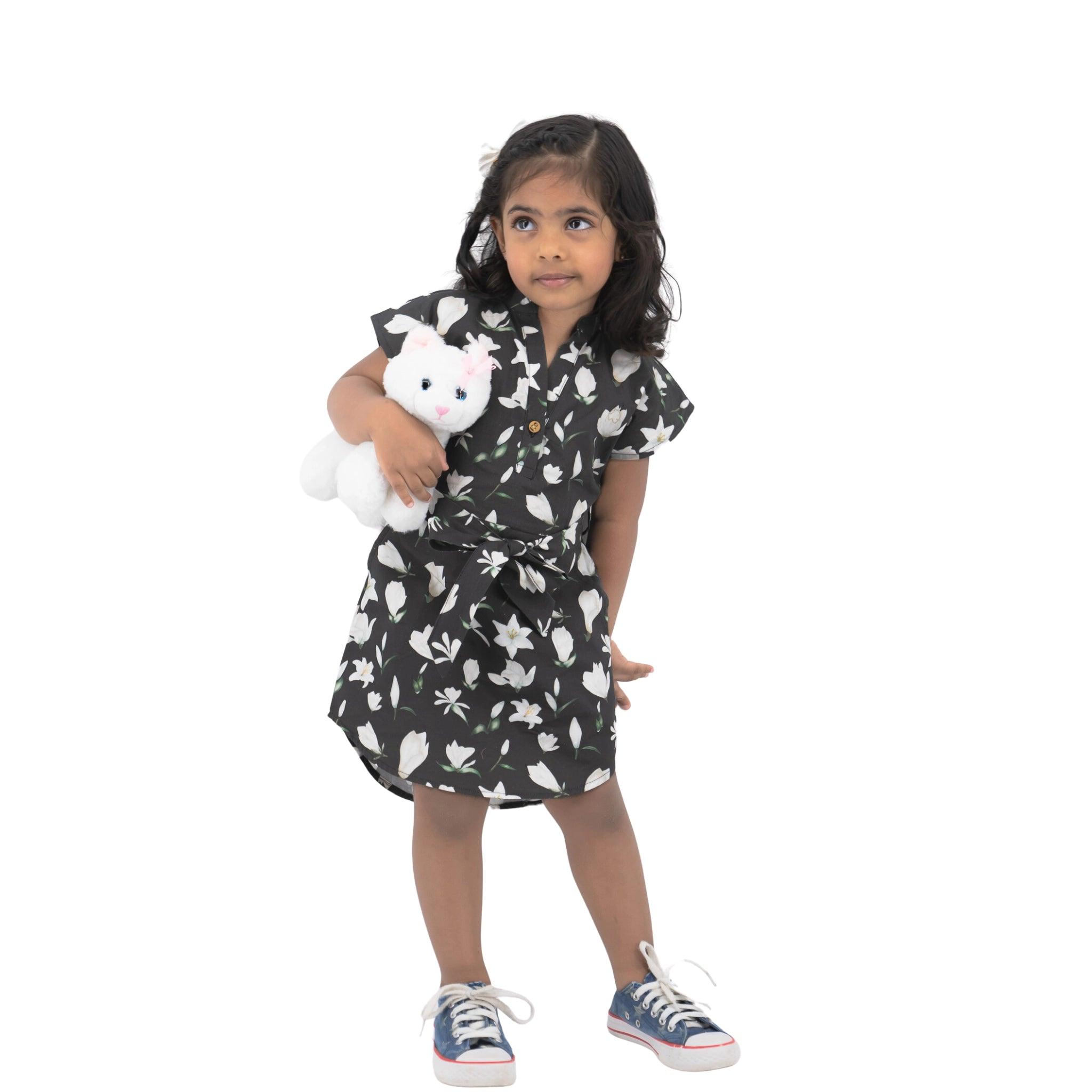 Young girl wearing a Karee Black Lilly Blossom Cotton Shirt Dress and sneakers, holding a white stuffed toy, standing against a white background.