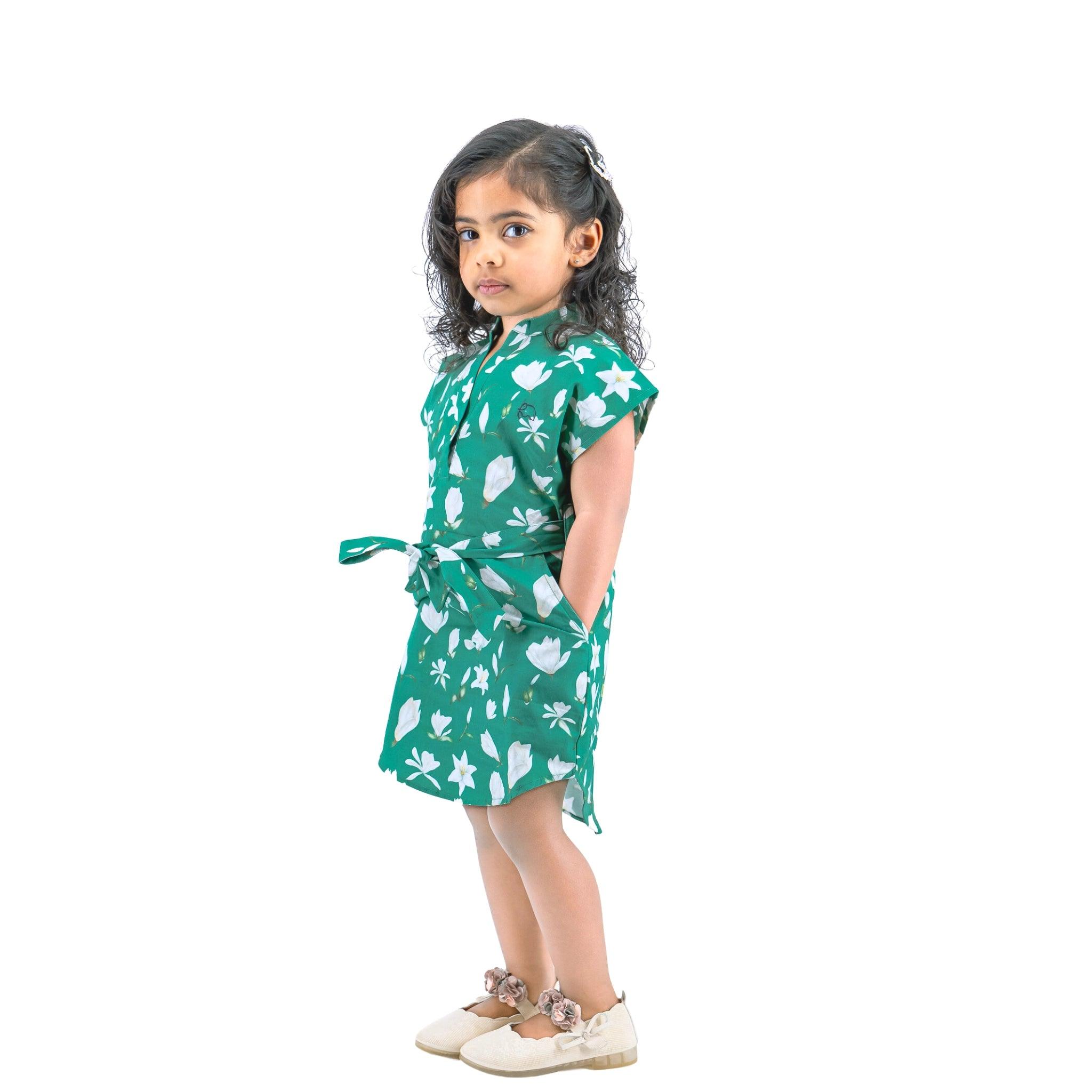 A young girl with curly hair, wearing a Karee Bottle Green Lilly Blossom Cotton Shirt Dress for Kids and white shoes, stands looking over her shoulder against a white background.
