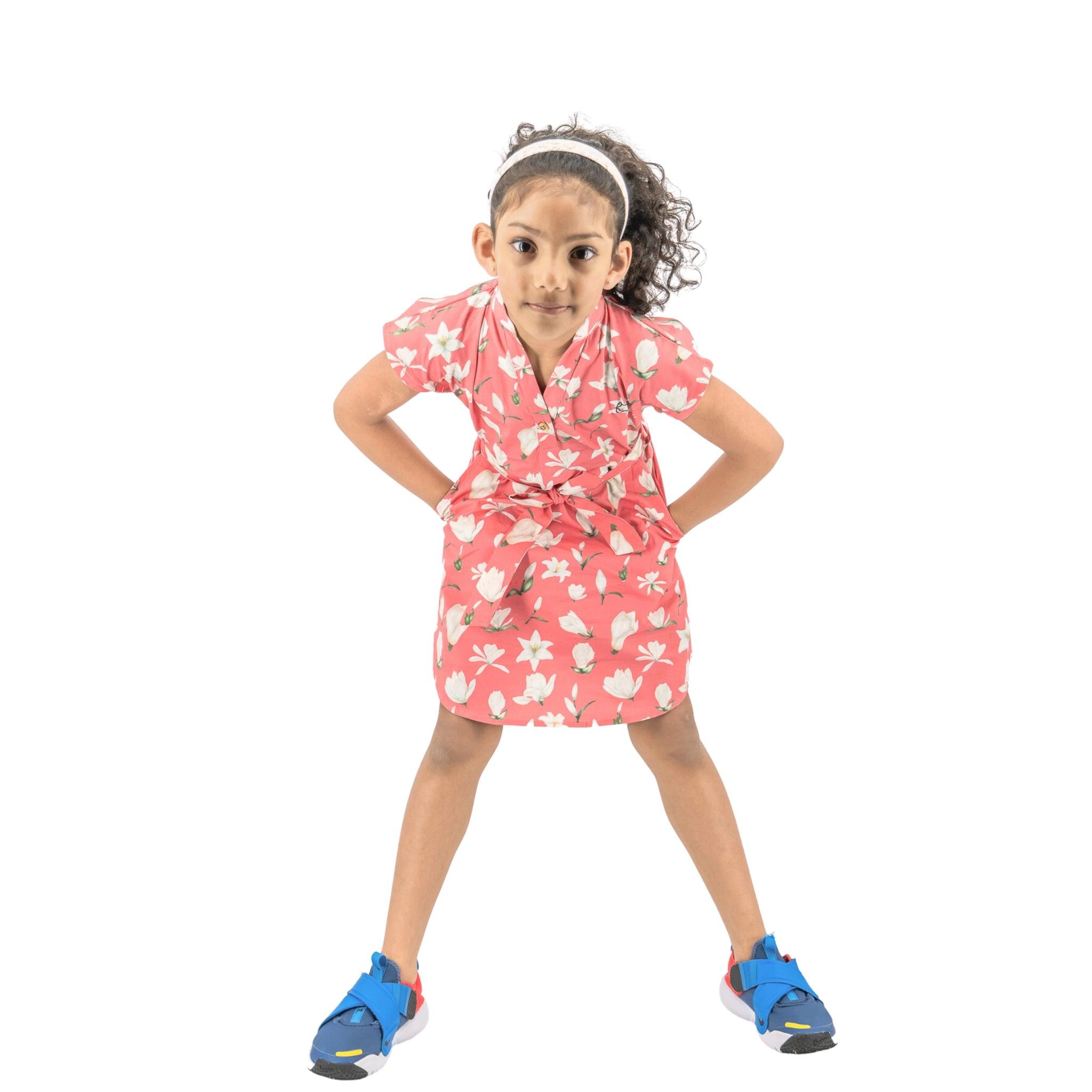 A young girl standing with hands on hips, wearing a Karee Mineral Red Lilly Blossom Cotton Shirt Dress and blue sneakers, looking at the camera with a slight frown on a white background.