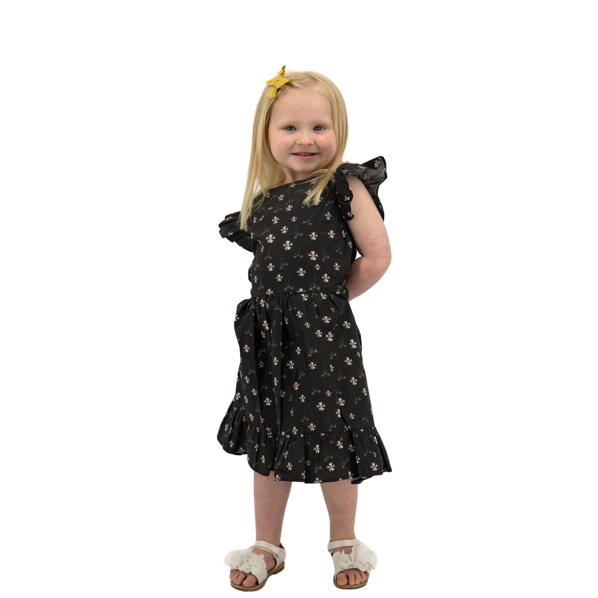 Young girl in a Karee Petite Blossom Black Cotton Dress and yellow hair bow standing against a white background, smiling.