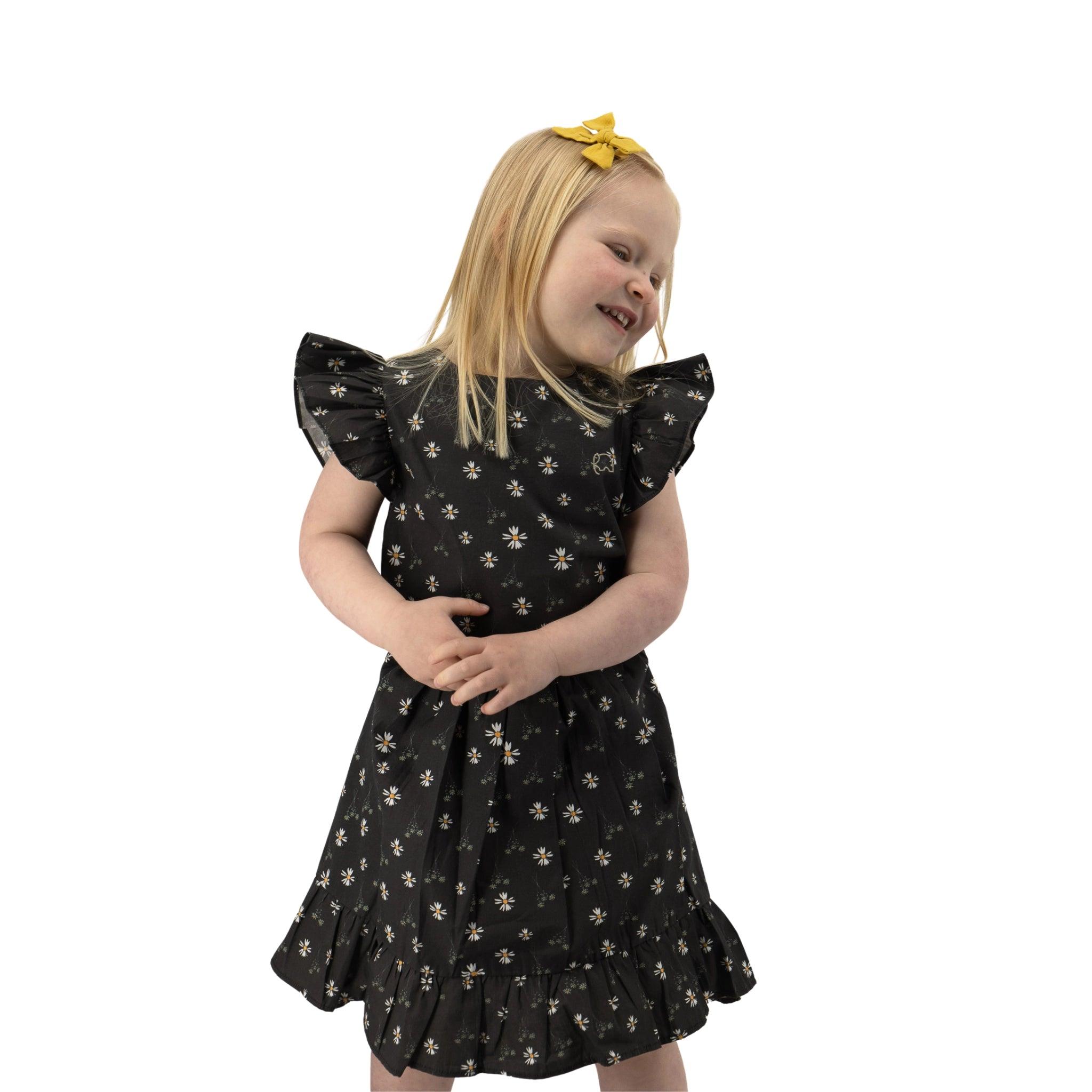 A young girl with a yellow hair bow and a Karee Petite Blossom Black Cotton Dress smiling and looking to her right, standing against a white background.