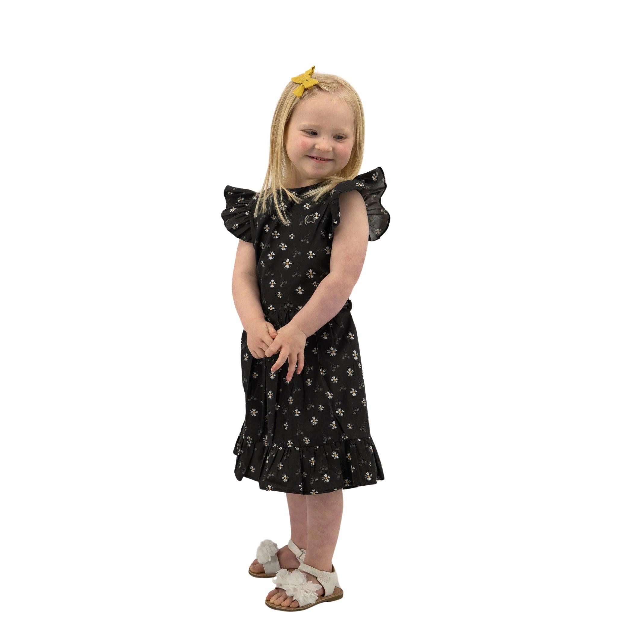 Young girl with blonde hair wearing a Karee Petite Blossom Black Cotton Dress with floral patterns and white sandals, posing with her hands together and smiling.