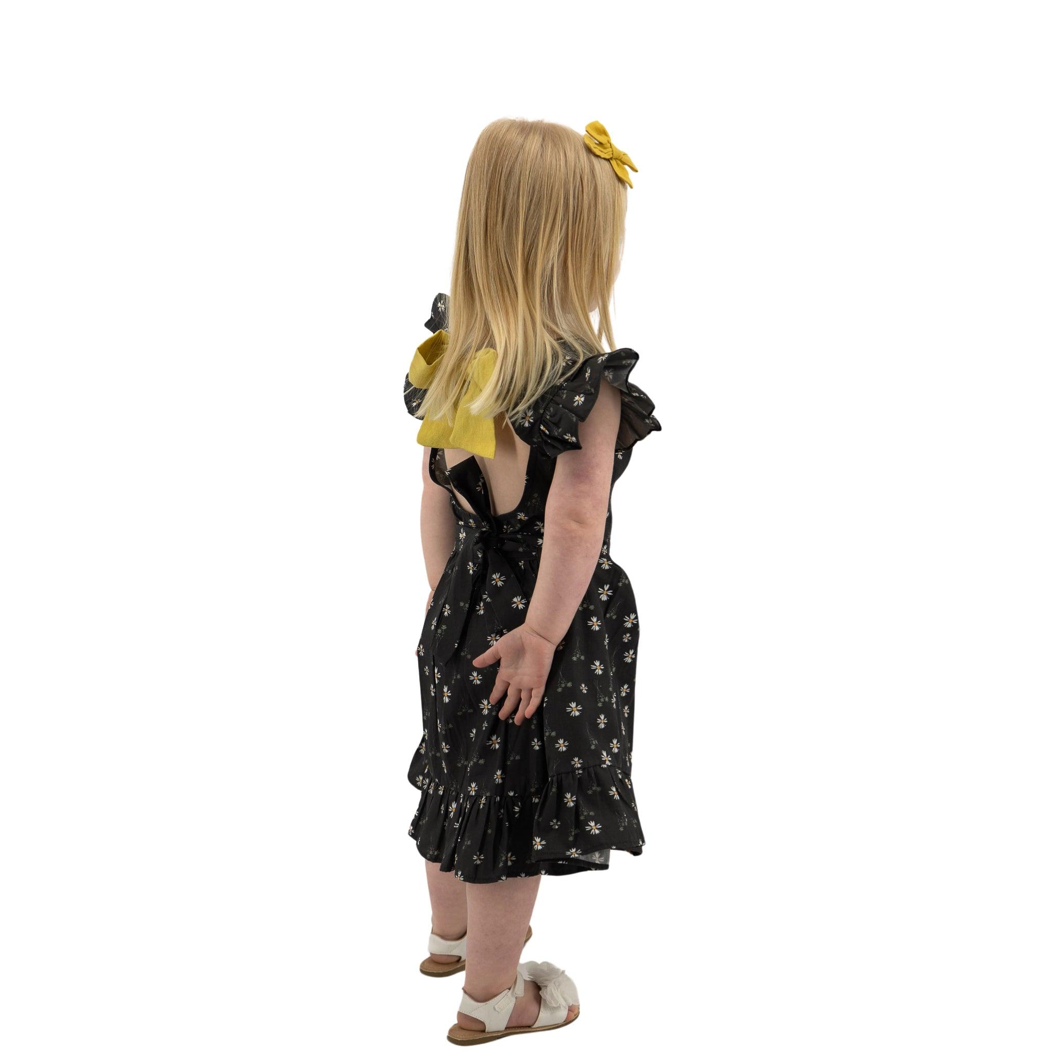 Young girl with blonde hair wearing a Karee Petite Blossom Black Cotton Dress and white shoes, standing in profile against a white background.