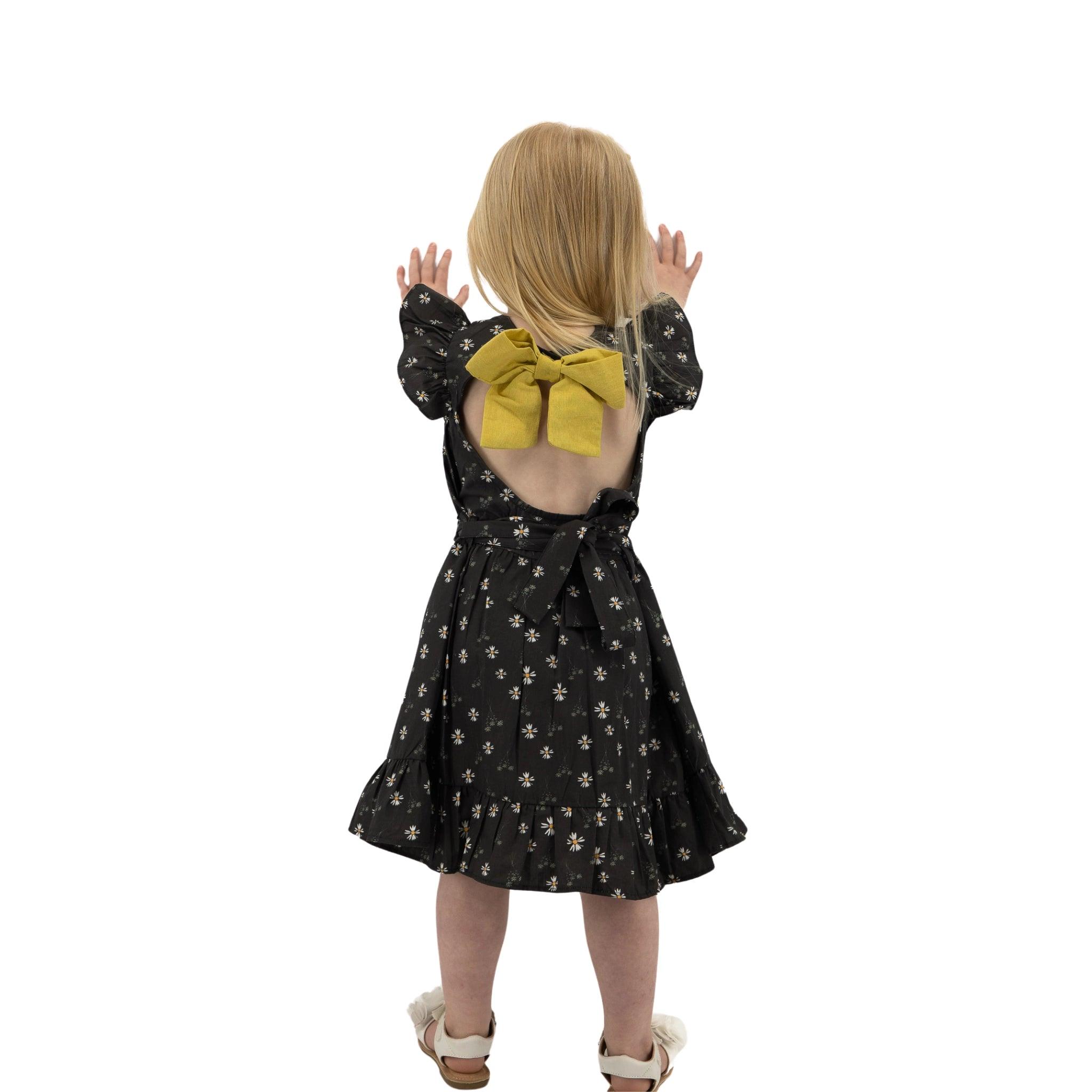 A young girl from behind, wearing a Karee Petite Blossom Black Cotton Dress with a large yellow bow, pressing her hands against an invisible surface.