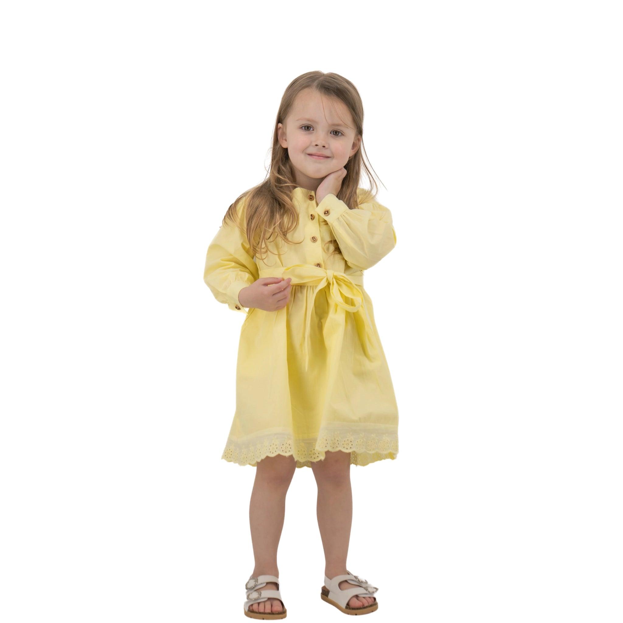 A young girl with long hair, wearing a Karee yellow long puff sleeve cotton dress and white sandals, stands smiling against a white background.