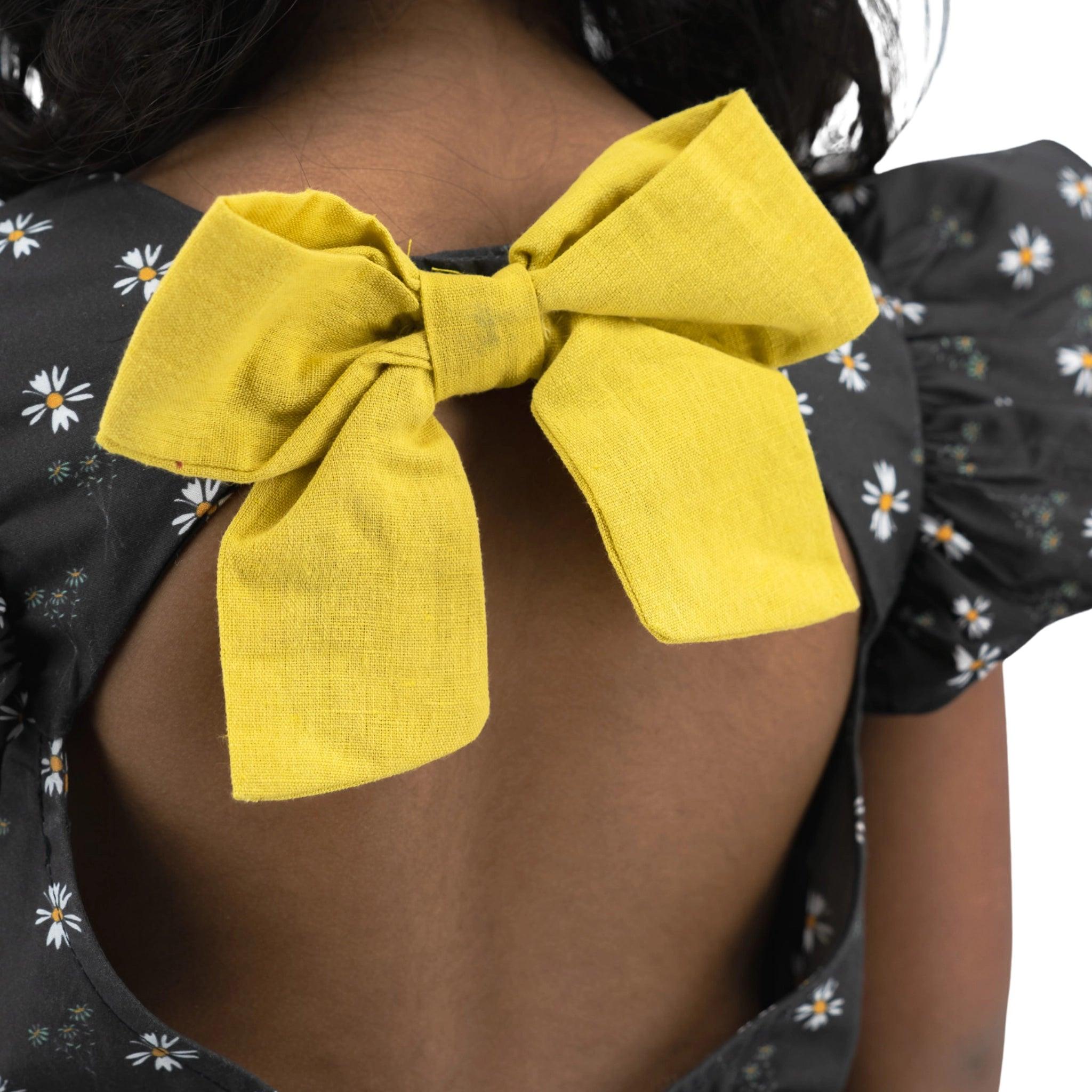 Close-up of a yellow bow tied at the back of a woman’s Petite Blossom Black Cotton Dress by Karee, focusing on the bow detail with part of her hair visible.