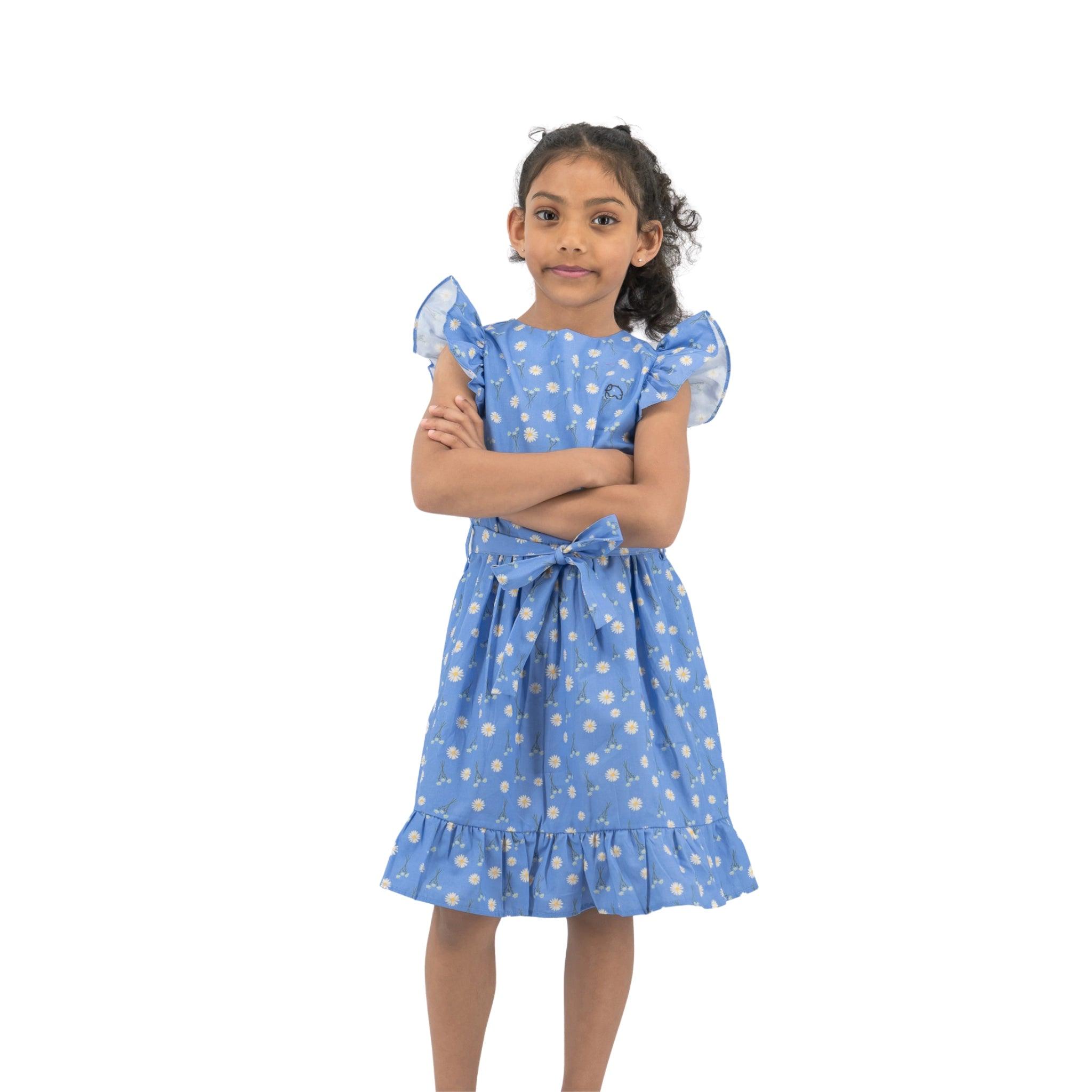 A young girl in a Karee Petite Blossom Cotton Dress in Corn Flour Blue standing with her arms crossed against a white background.