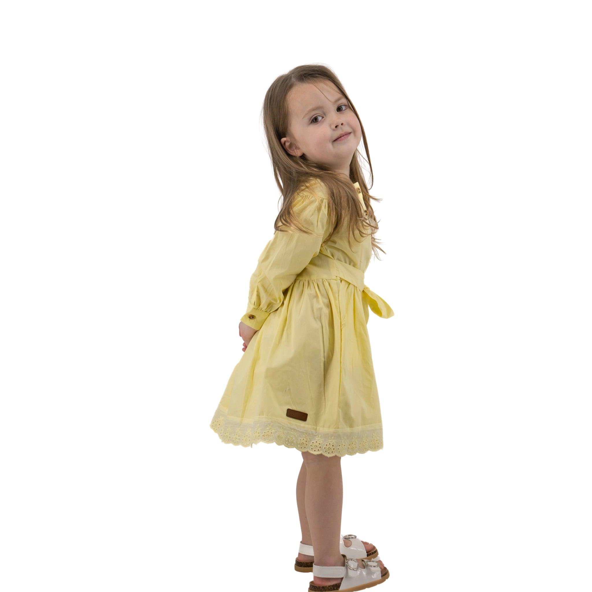 Young girl in a Karee yellow long puff sleeve cotton dress standing sideways and looking over her shoulder with a playful smile, isolated on a white background.