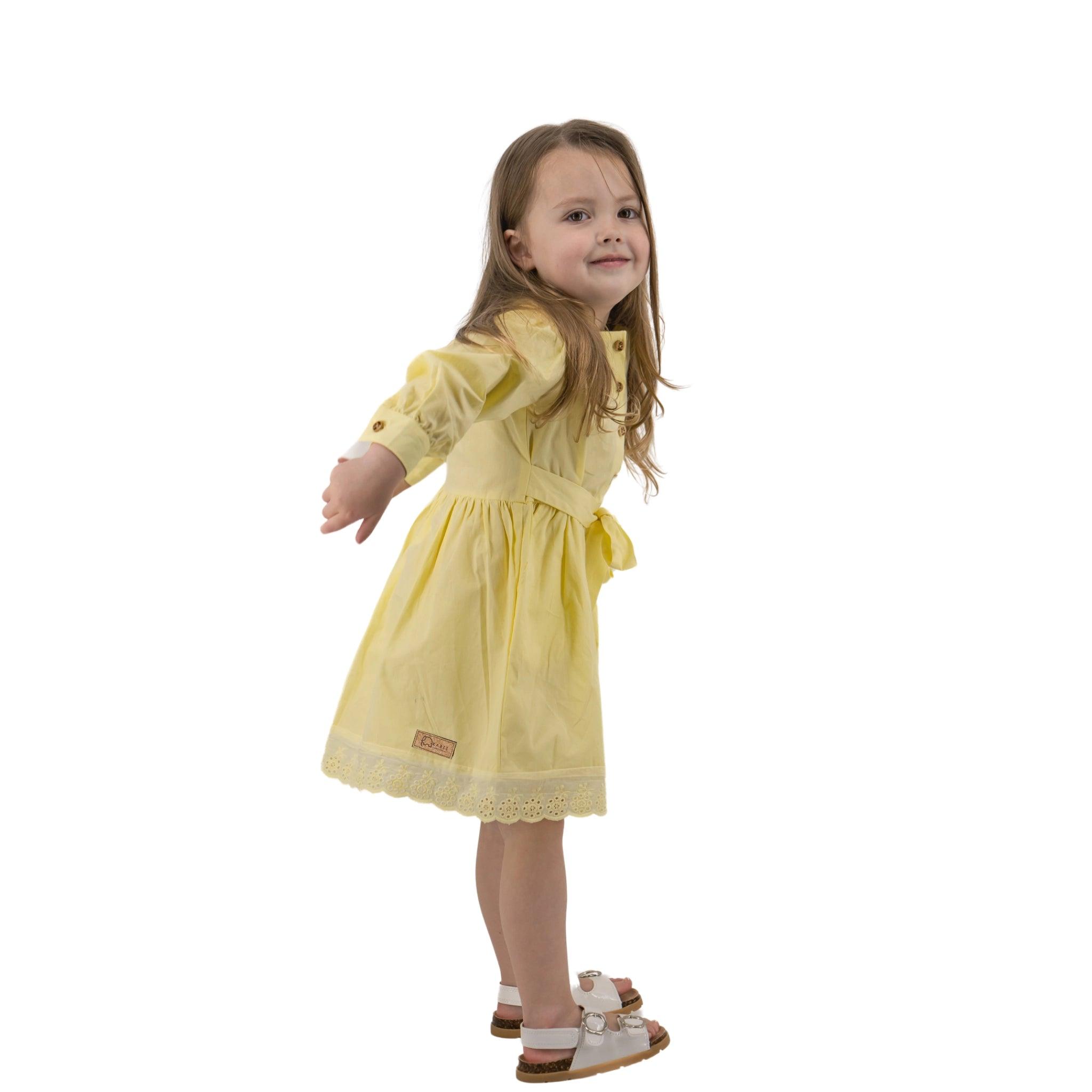A young girl in a Karee yellow long puff sleeve cotton dress and white sandals, standing with arms outstretched, smiling at the camera. White background.
