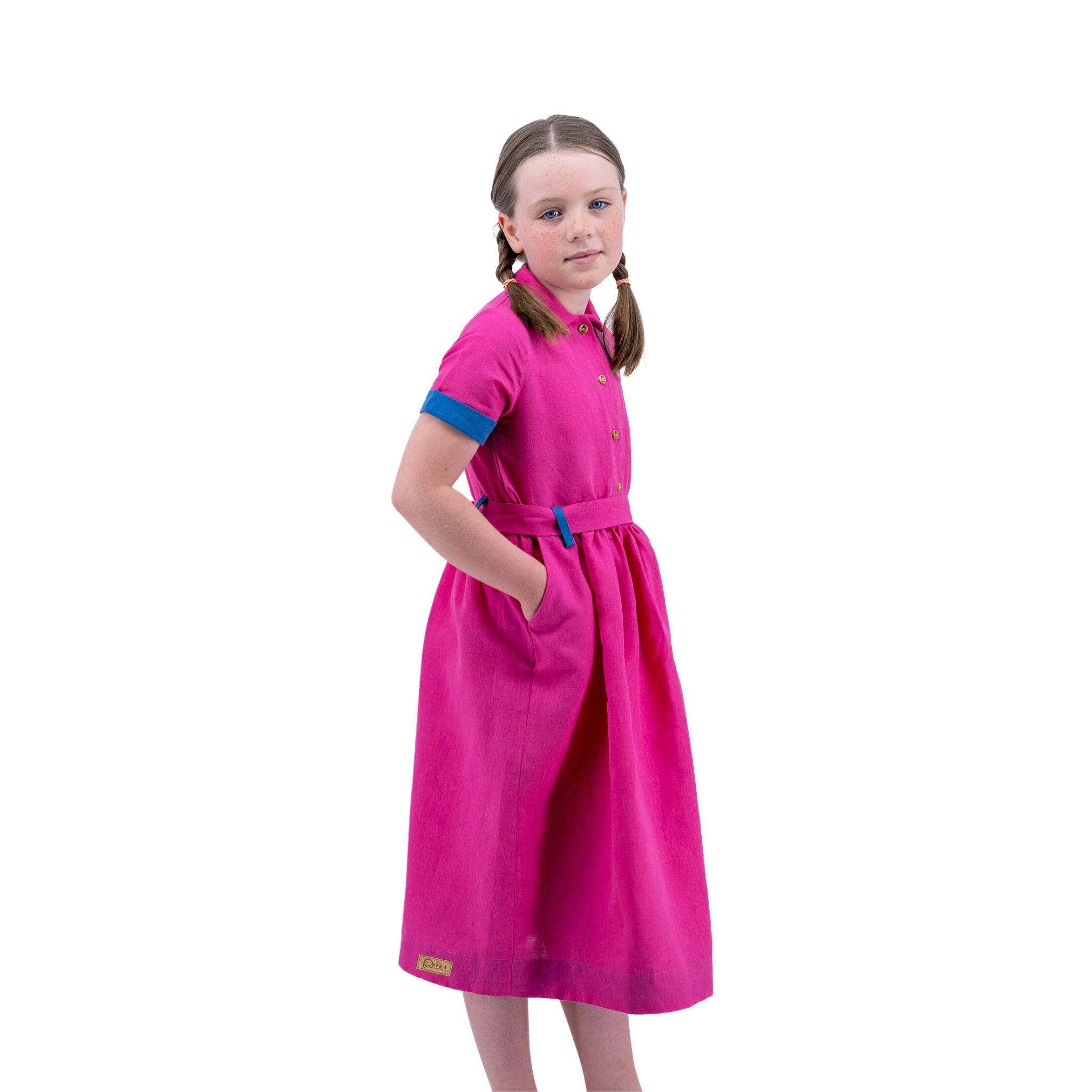 Young girl in a Karee fuchsia purple linen dress and braided hair, looking over her shoulder, standing against a white background.