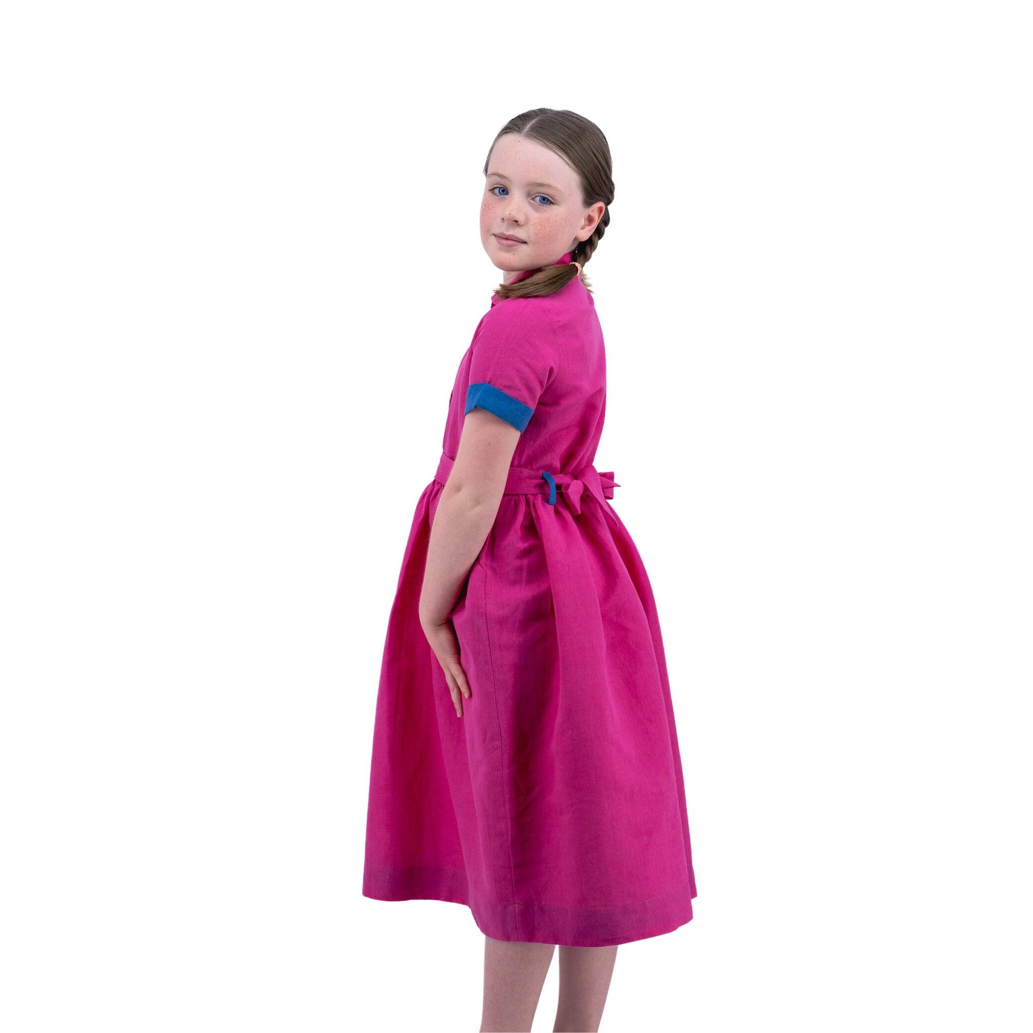 Young girl in a Karee fuchsia purple linen dress for girls with blue trim, standing sideways and looking over her shoulder against a white background.