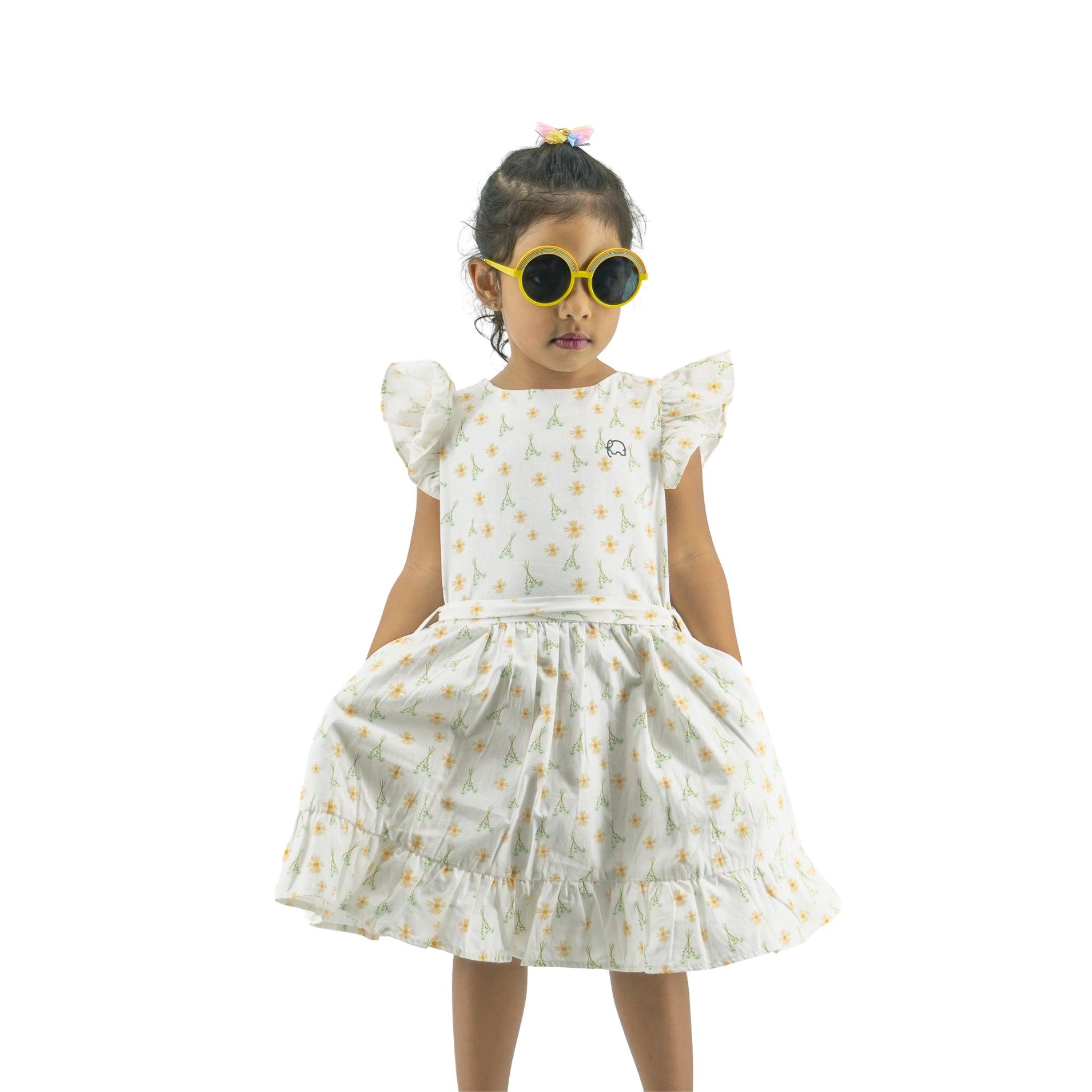 Young girl in a Karee Petite Blossom Cotton Dress in Smoked Pearl with star patterns, wearing large yellow sunglasses, standing against a white background.