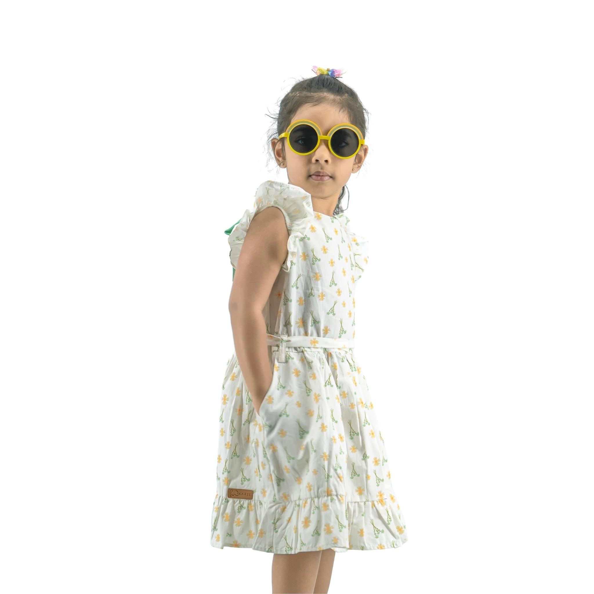 Young girl wearing sunglasses and a Karee Petite Blossom Cotton Dress in Smoked Pearl, looking over her shoulder, isolated on a white background.