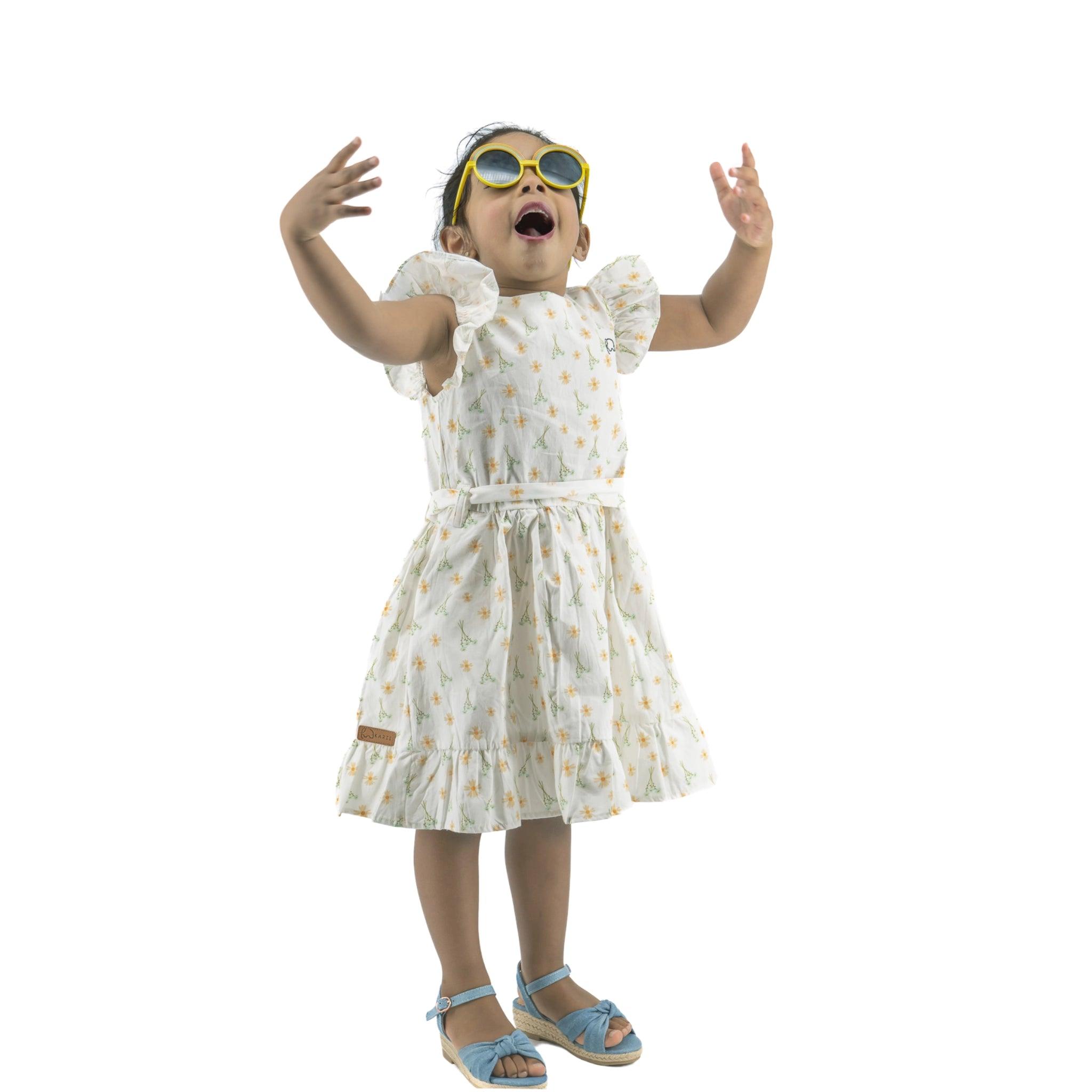 A young girl wearing a Karee Petite Blossom Cotton Dress in Smoked Pearl, sandals, and oversized yellow goggles, joyfully raises her arms, isolated on a white background.