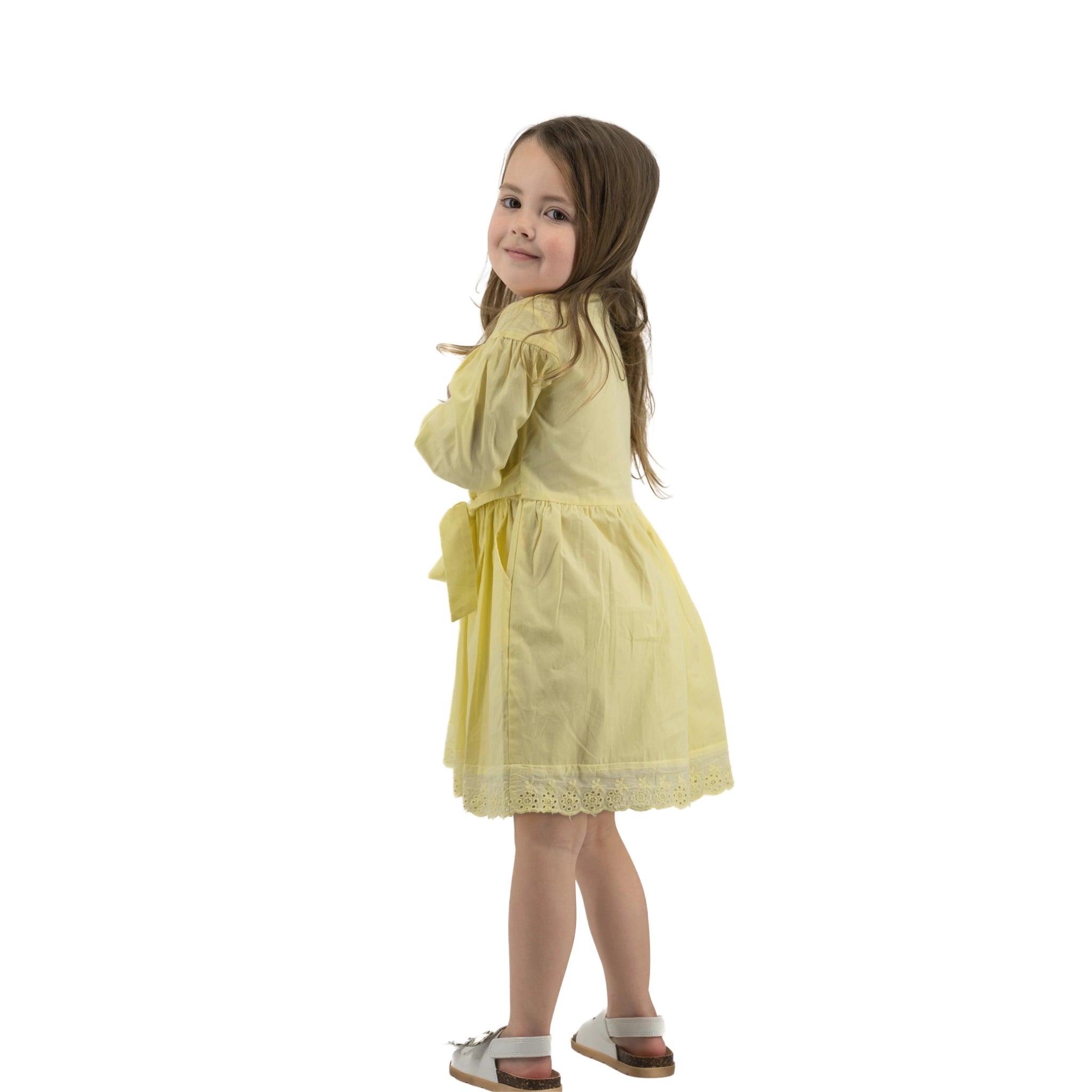 Young girl in a Karee yellow long puff sleeve cotton dress smiling over her shoulder, standing against a white background.