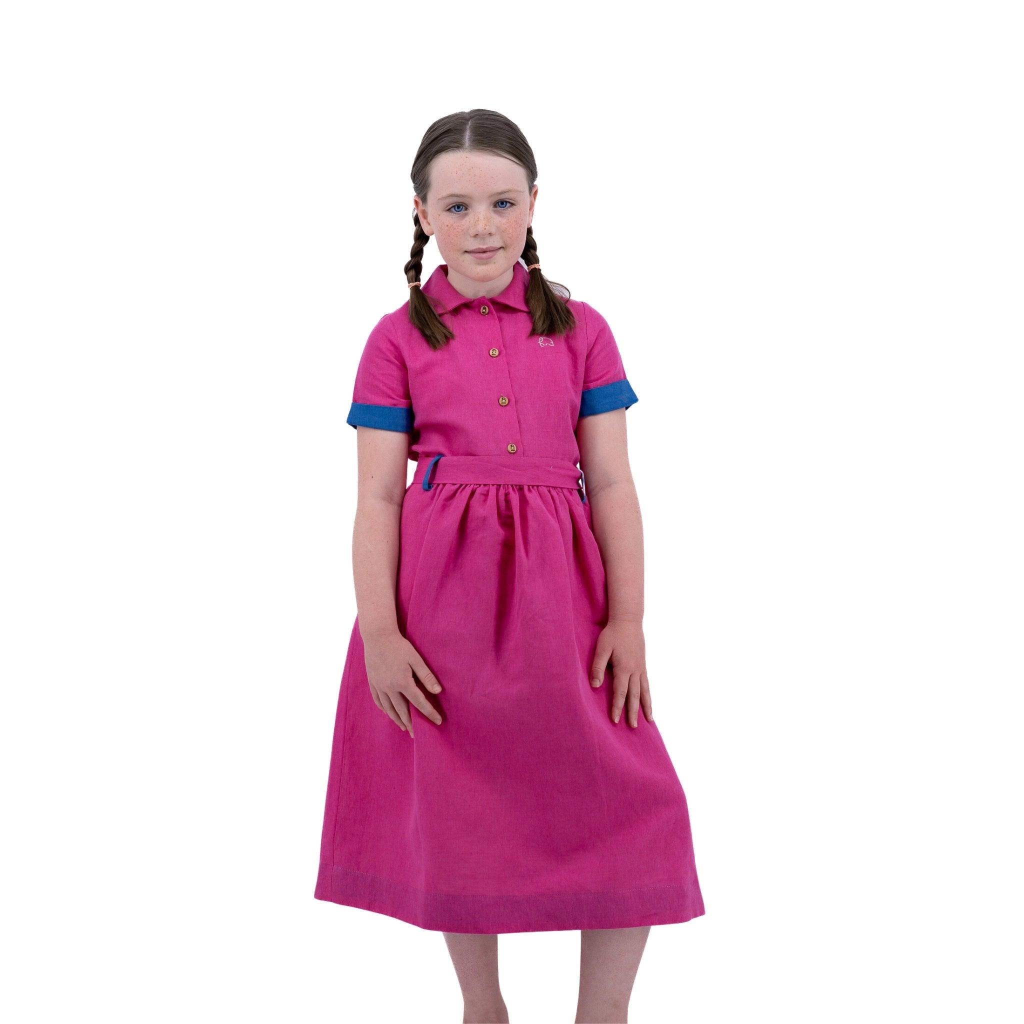 Young girl in a Karee fuchsia purple linen dress for girls with blue cuffs, standing with hands on hips and pigtails, isolated on a white background.