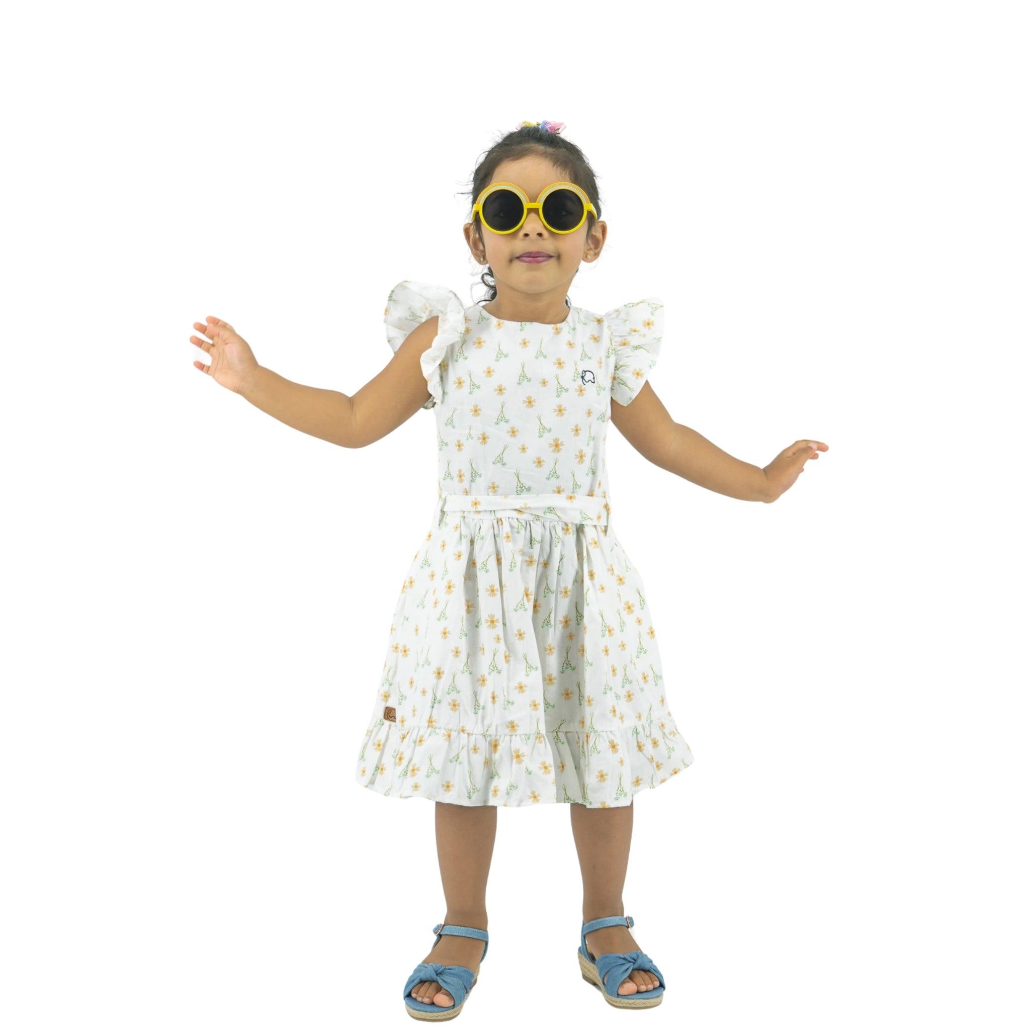 A young girl wearing a Karee Petite Blossom Cotton Dress in Smoked Pearl and sunglasses stands with her arms outstretched, isolated on a white background.