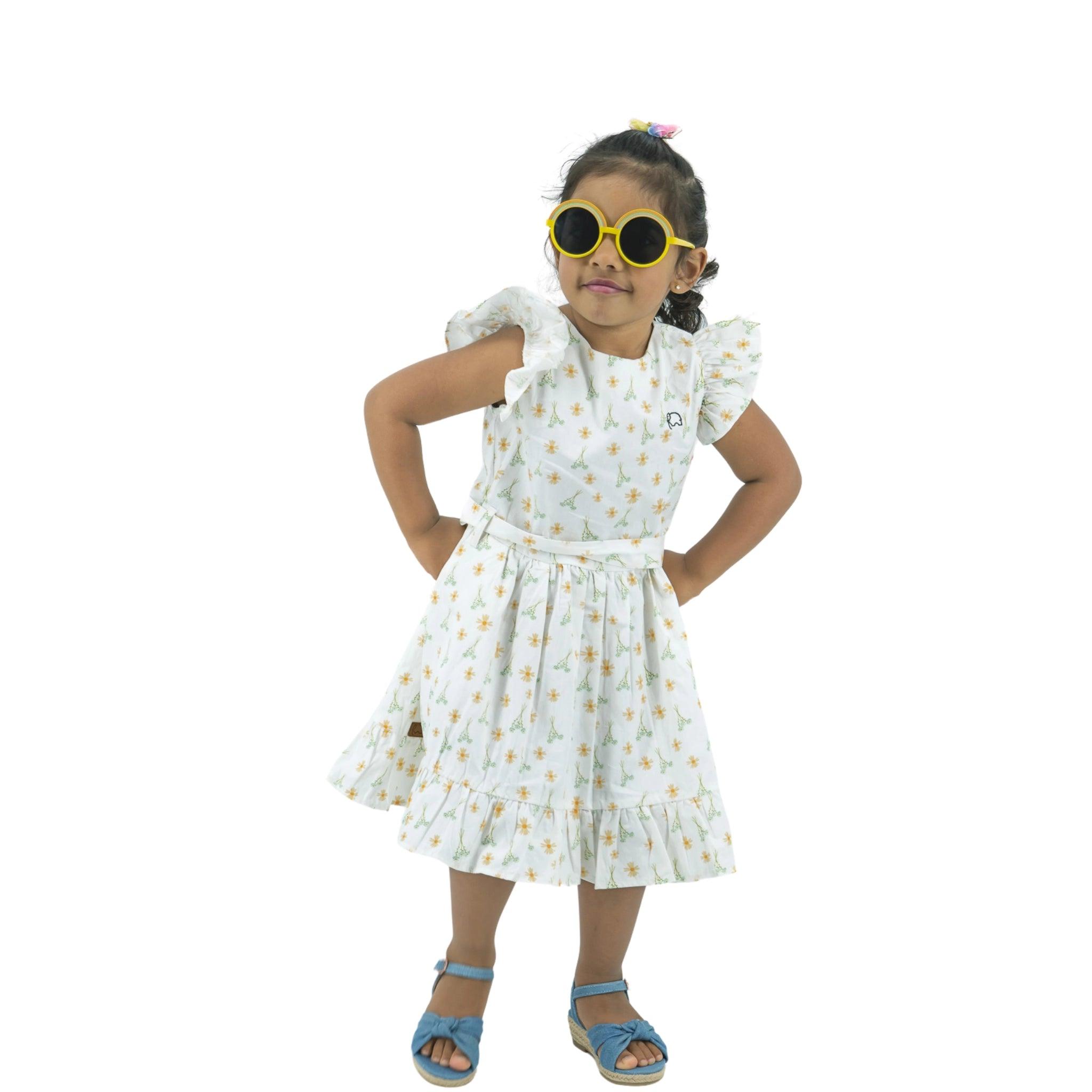 A young girl stands with hands on her hips, wearing a Karee Petite Blossom Cotton Dress in Smoked Pearl, yellow sunglasses, and blue sandals, against a white background.