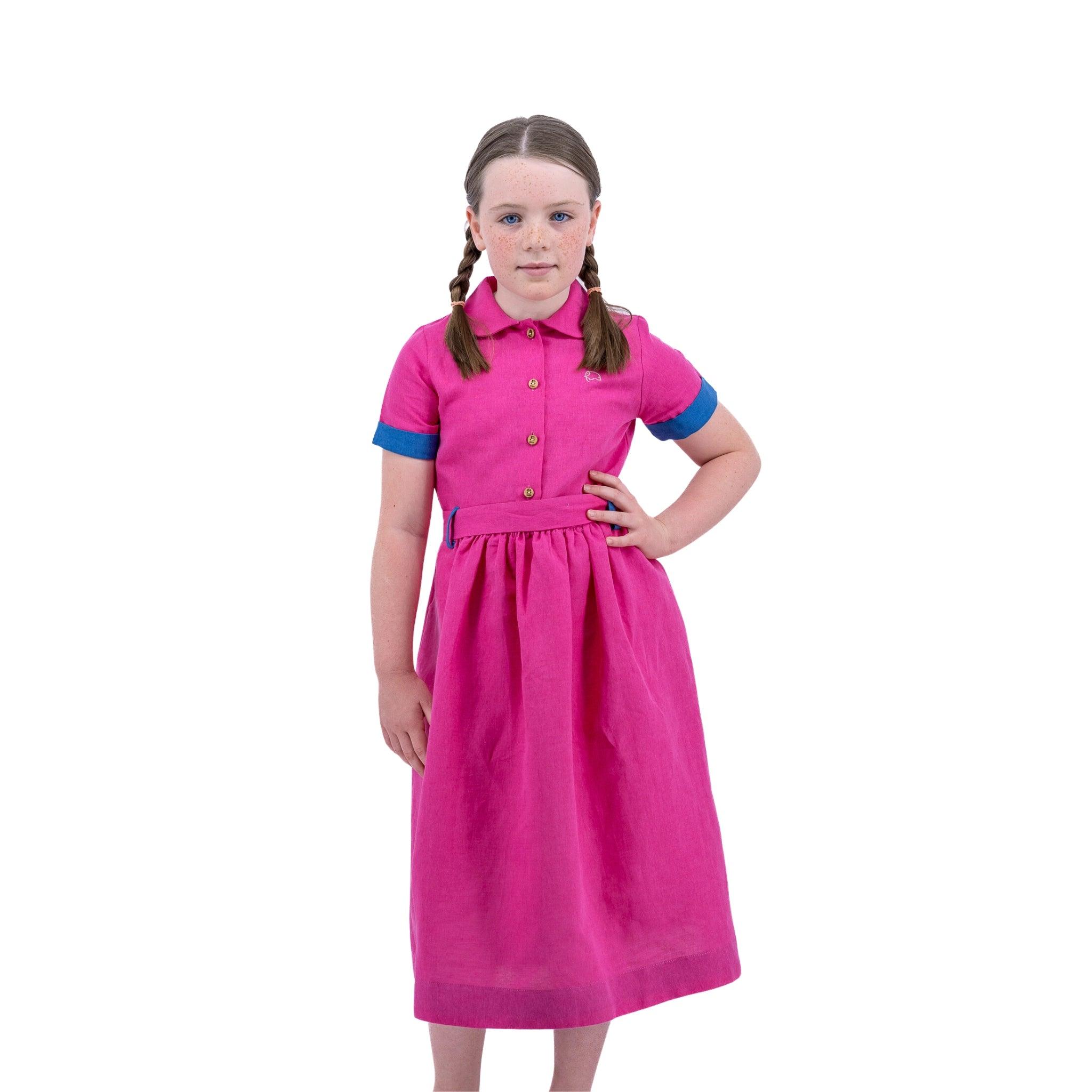 Young girl in a Karee fuchsia purple linen dress with blue sleeves, standing with hands on hips against a white background.