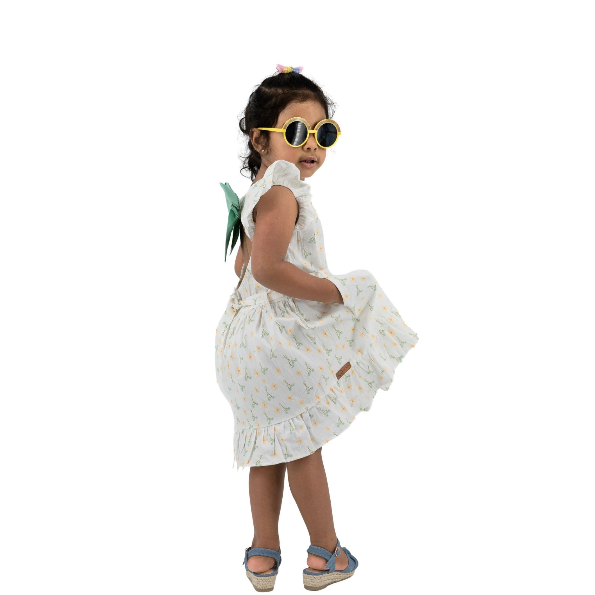 Young girl in a Karee Petite Blossom Cotton Dress in Smoked Pearl and yellow sunglasses turning playfully, isolated on white background.
